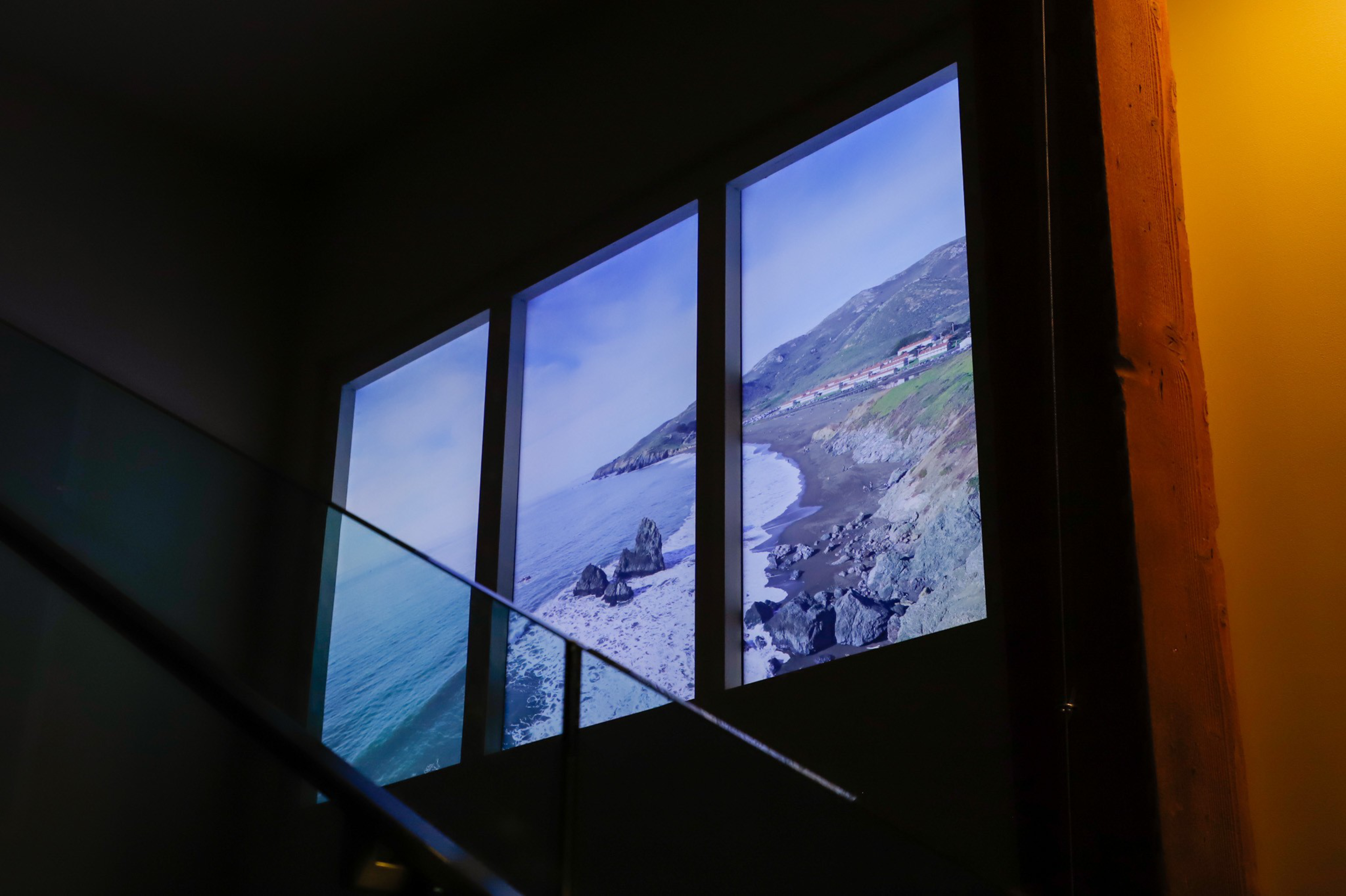 Three vertical windows display a coastal scene with rocky cliffs, the ocean, and a clear sky, viewed from a dimly lit interior space.
