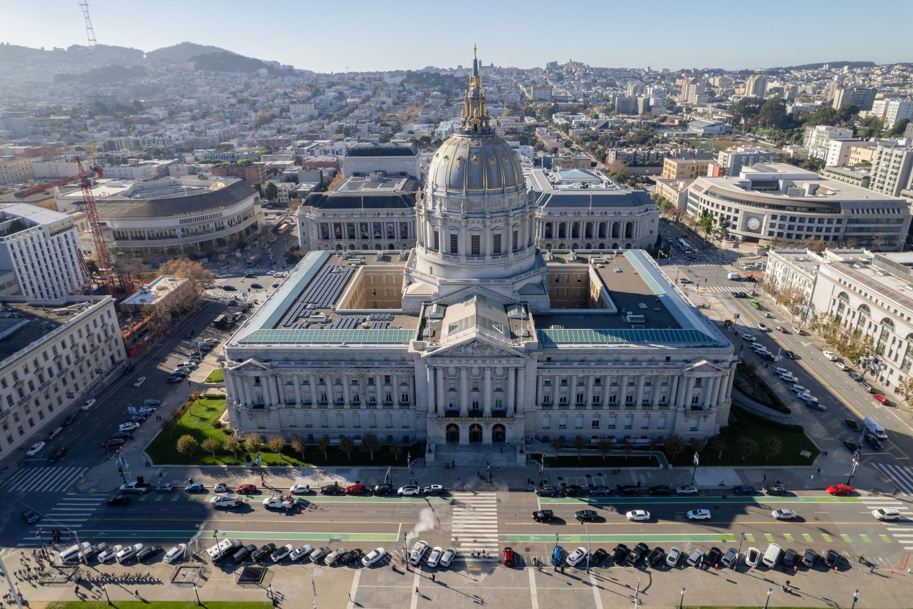 Aerial view of a large, ornate building with a dome, surrounded by streets and other buildings in a hilly urban area.