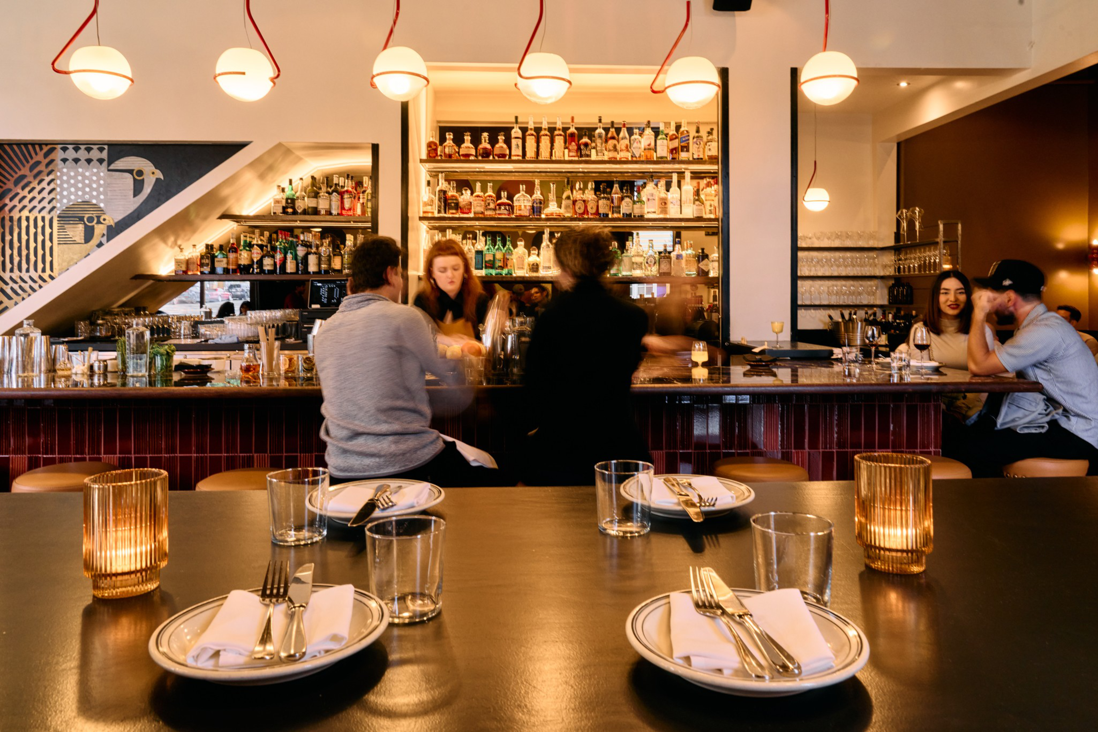 A cozy bar with patrons seated at the counter, shelves stocked with bottles, warm lighting, and a table set with plates, cutlery, and glasses in the foreground.