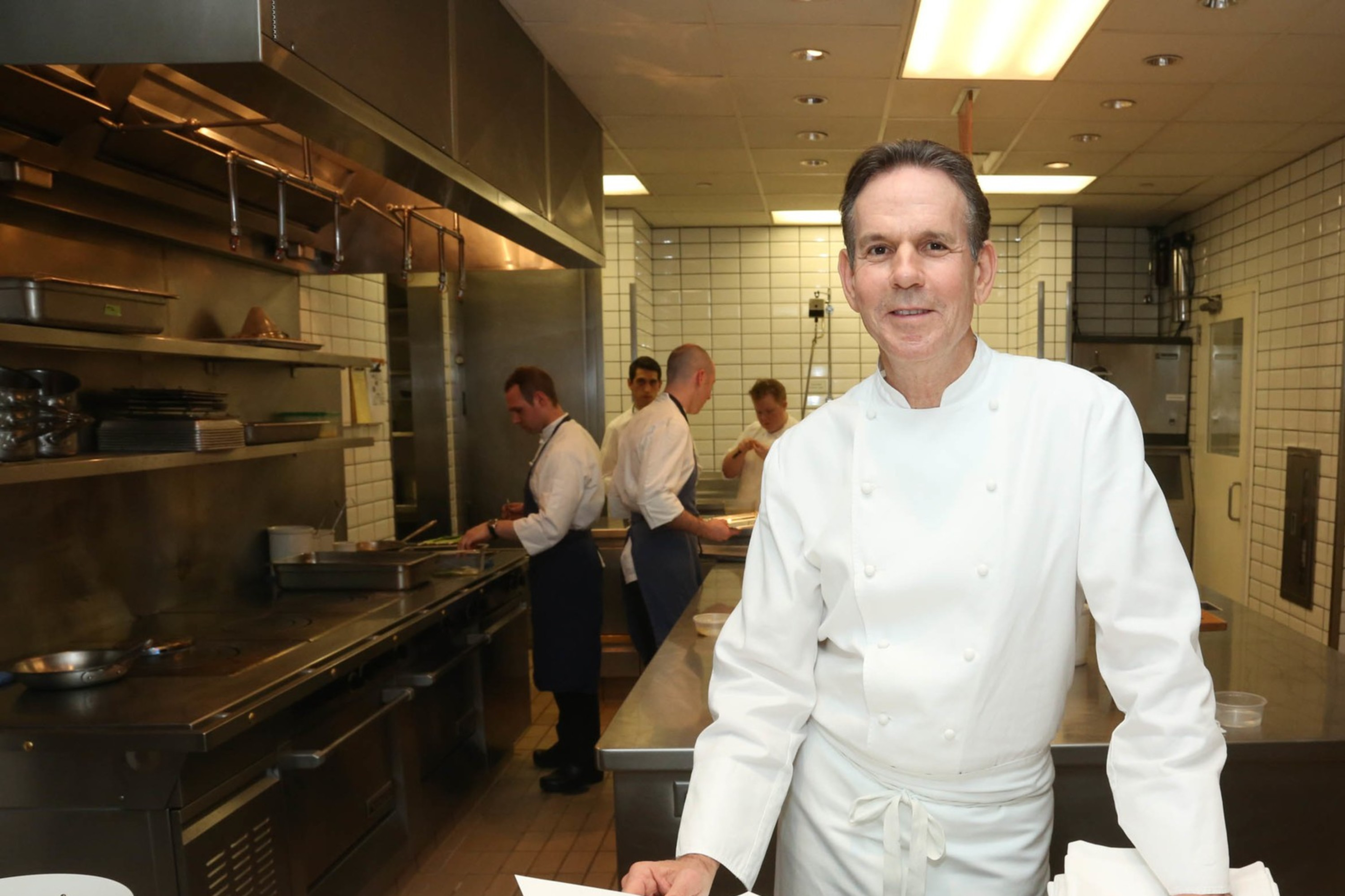 A chef in a white uniform stands in a spotless, tiled kitchen. He holds a paper, with a bowl of red petals nearby. Behind him, others work in the kitchen.