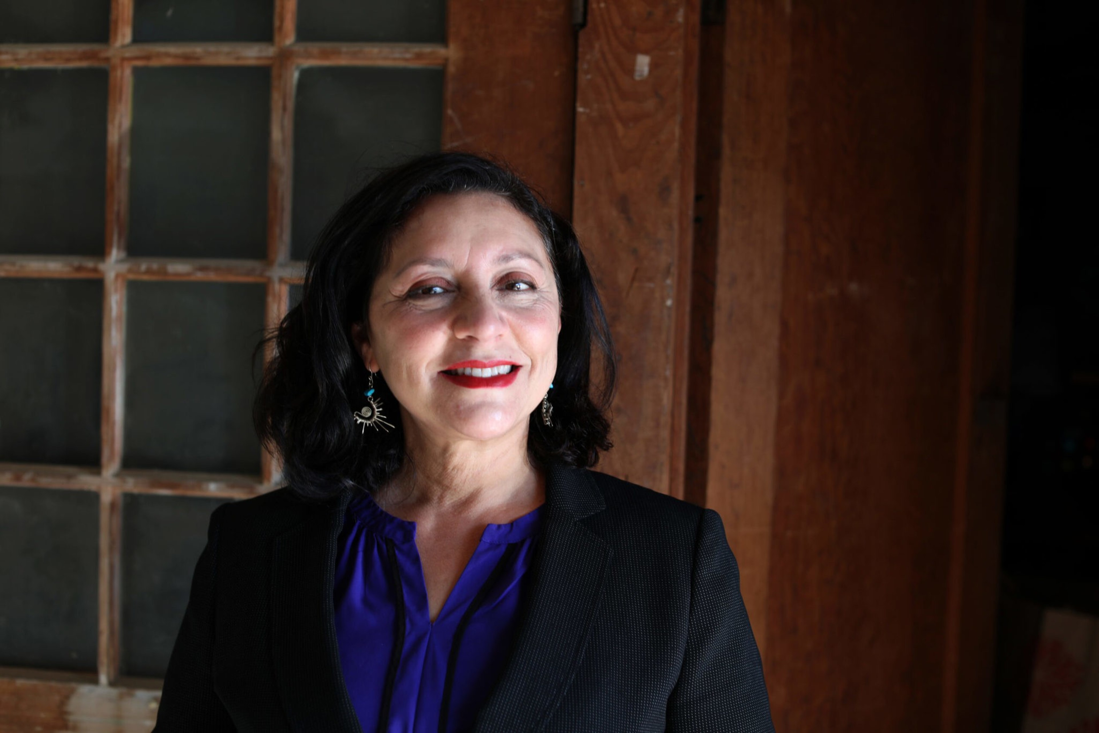 A woman with shoulder-length black hair, wearing a black blazer, blue blouse, and sun-shaped earrings smiles against a wooden and glass window background.