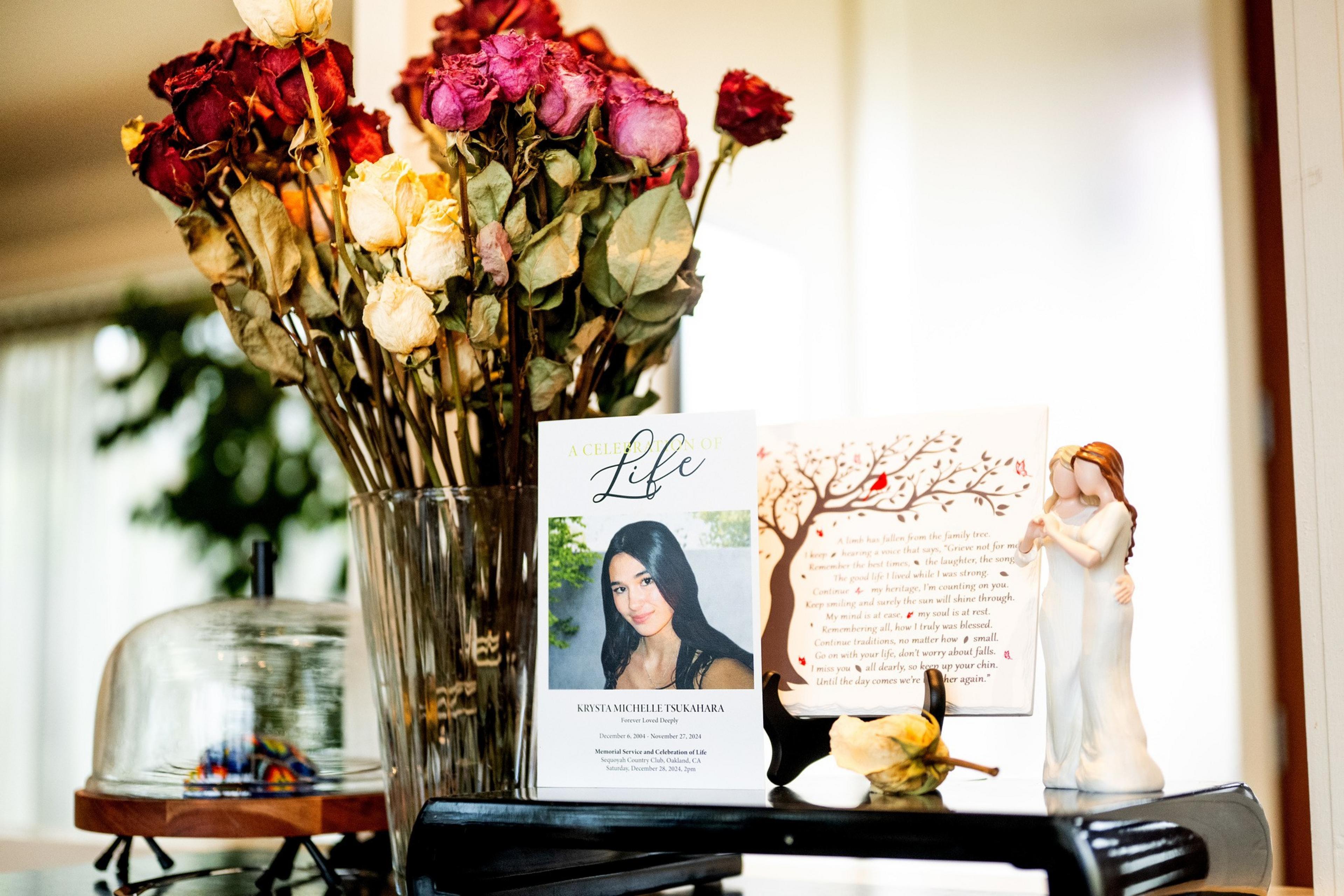 A vase of dried roses stands beside a memorial card with a woman's photo and a small statuette of two figures embracing, set on a table.