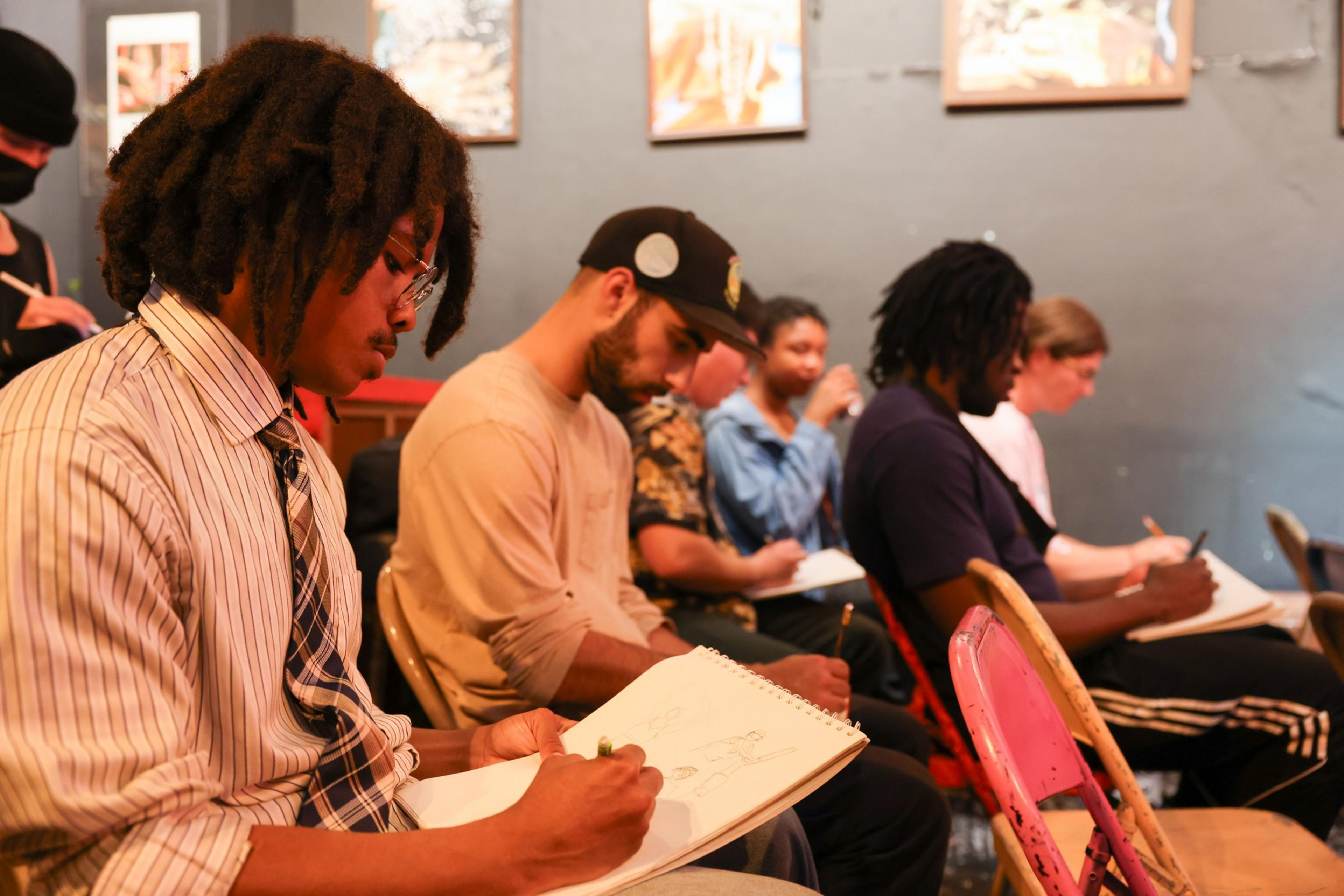 Five people sit in a row on folding chairs, focused on sketching or drawing in notebooks, in a classroom or workshop setting.