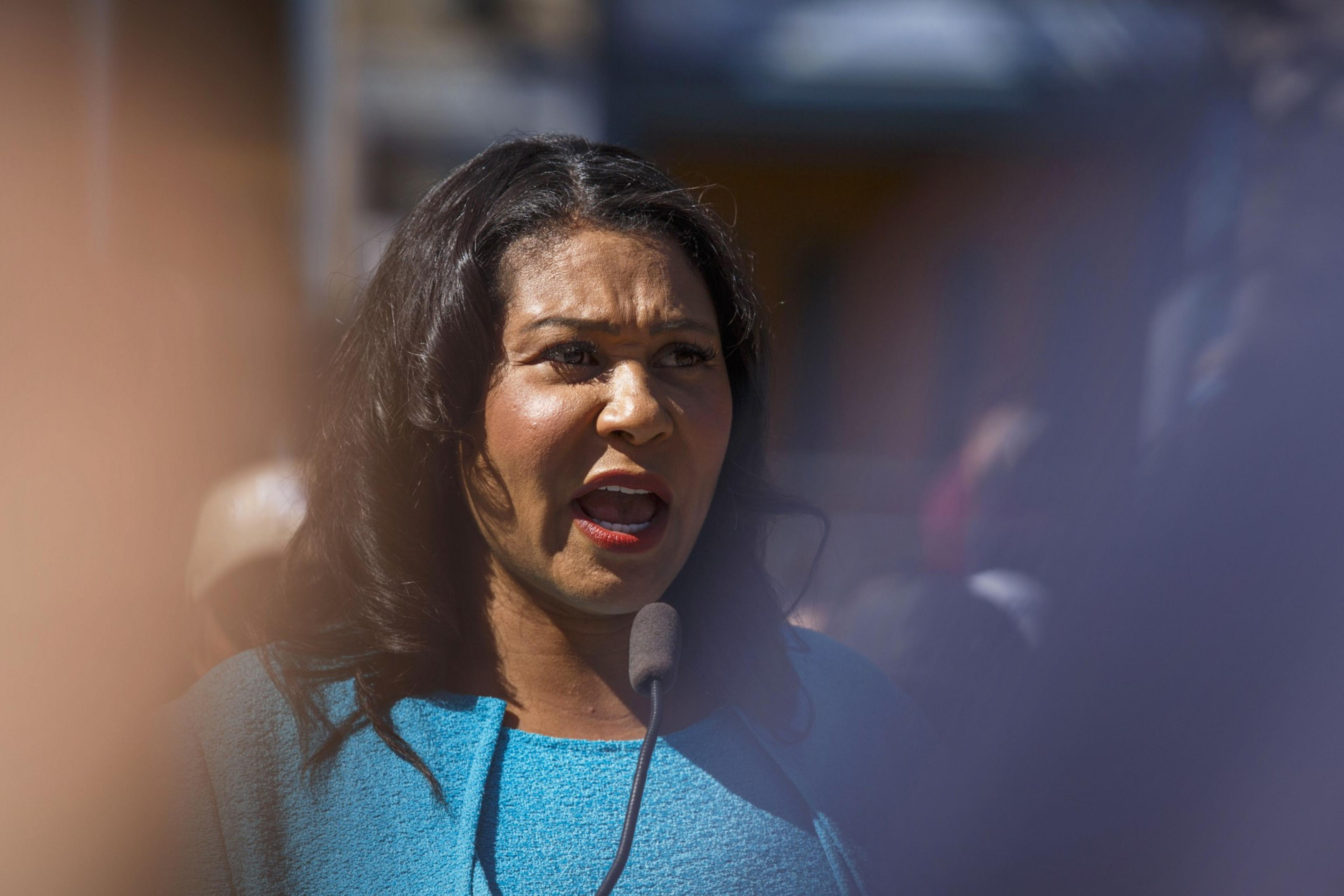 A person is speaking into a microphone, wearing a bright blue top, with a blurry background.