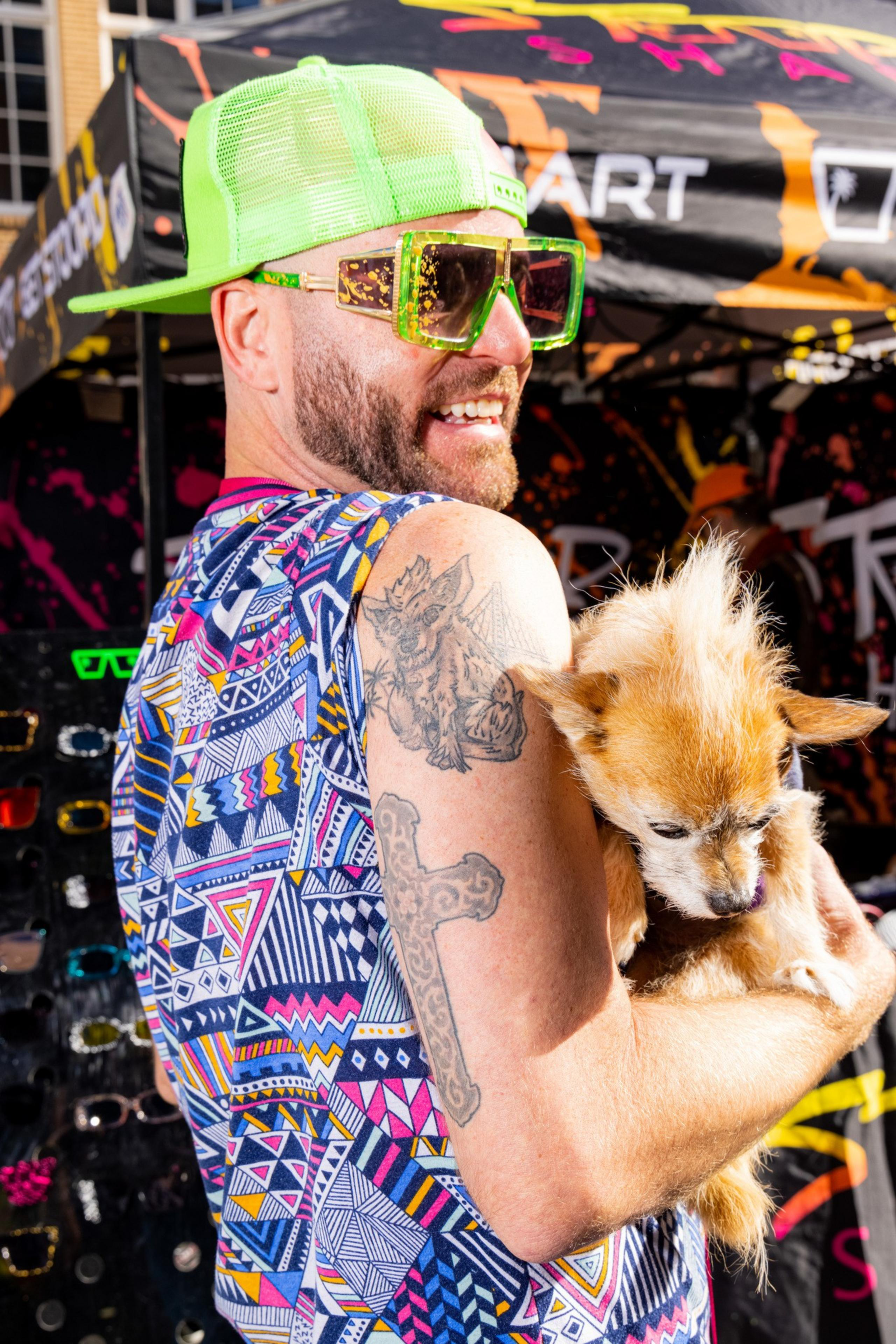 A man wearing a neon green hat, large colorful sunglasses, and a patterned shirt holds a small dog with spiky fur. He has tattoos on his arm.