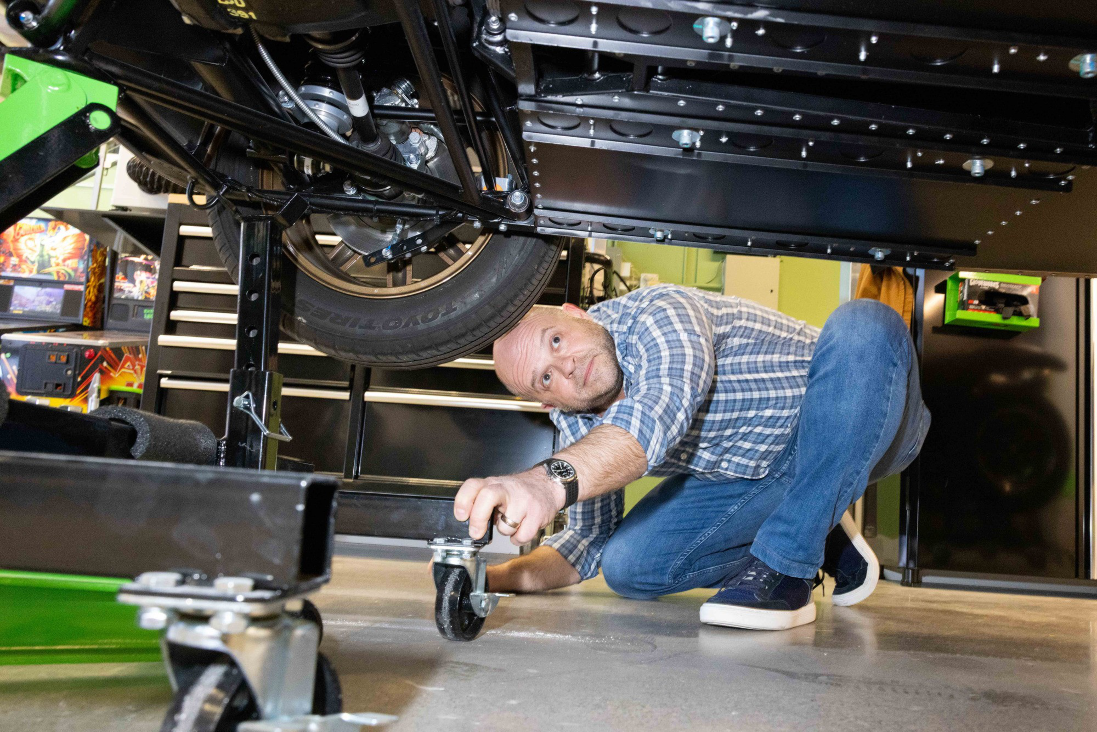 A person in a plaid shirt and jeans crouches under a vehicle, adjusting a caster wheel. The garage setting has tool cabinets and vibrant wall decorations.