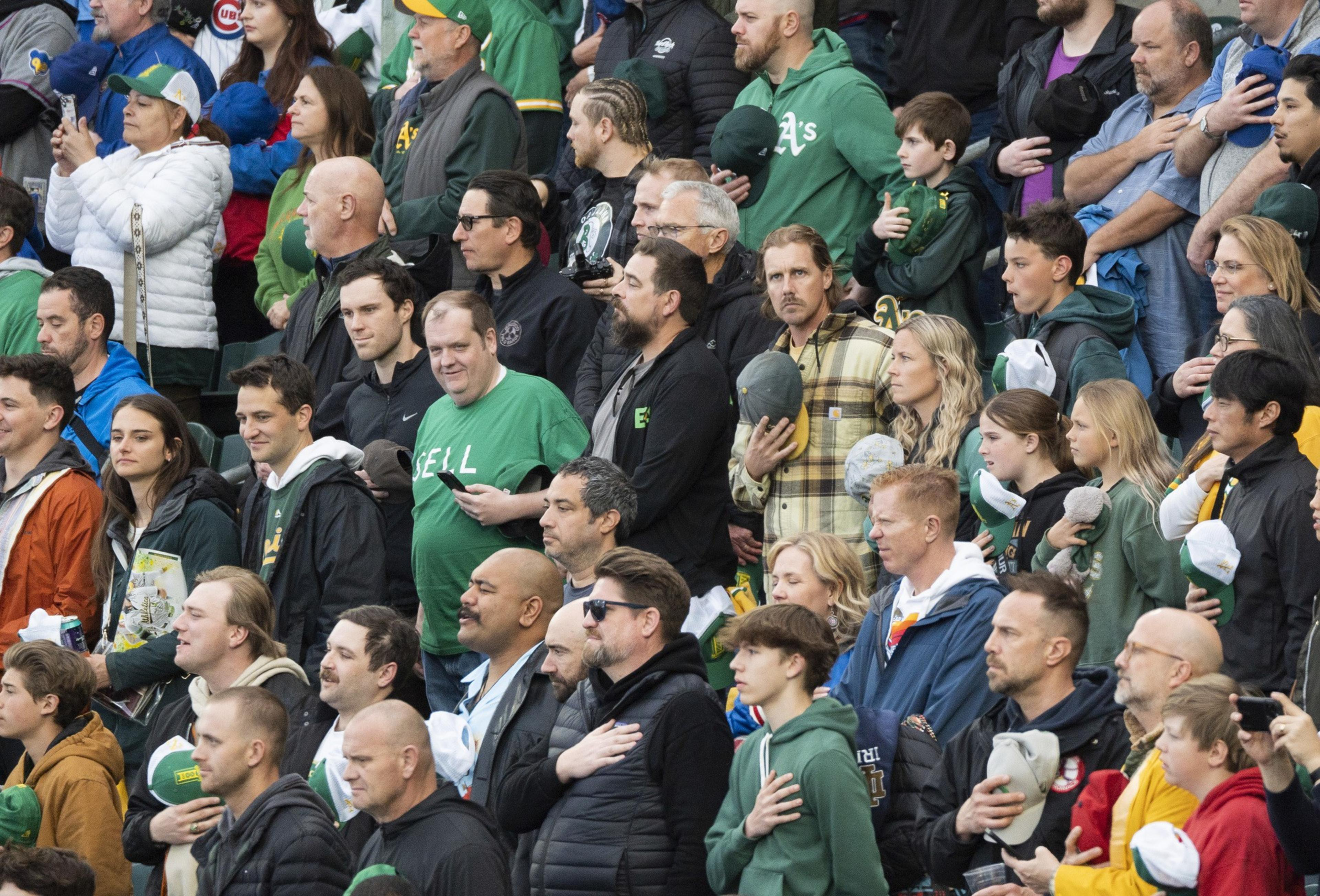 The image shows a crowd of people at a sports event, standing with hands over their hearts. They wear various team apparel, indicating a shared enthusiasm.