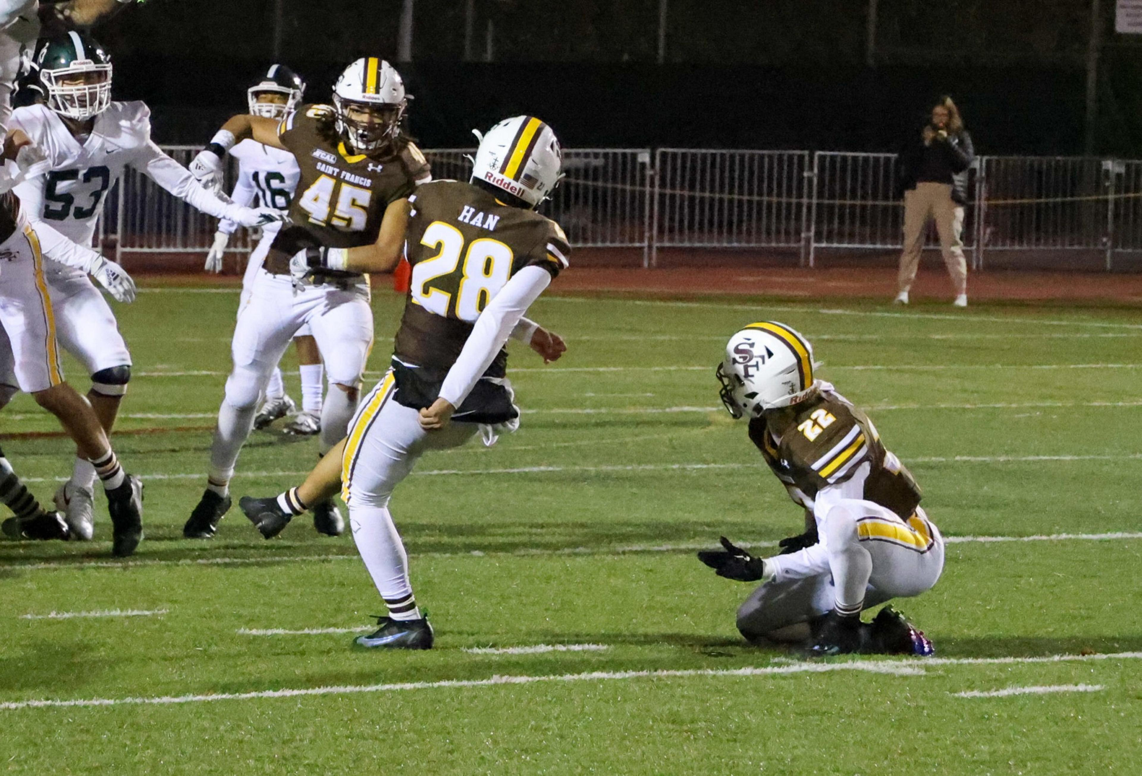 A football player in a brown jersey kicks the ball while a teammate crouches nearby, with opposing team players in white jerseys approaching.
