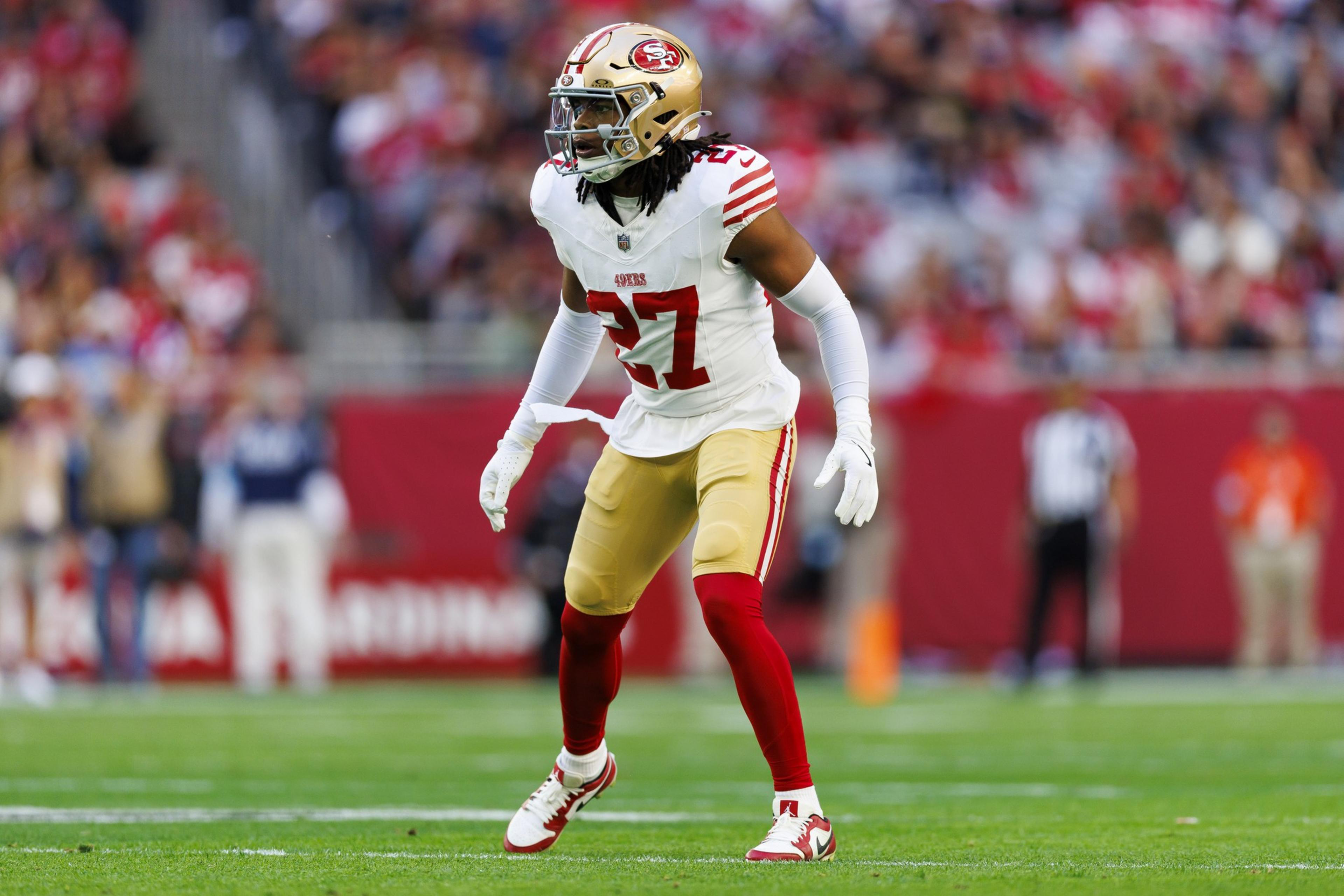 A football player wearing a white and red San Francisco 49ers uniform, number 27, stands ready on the field during a game.