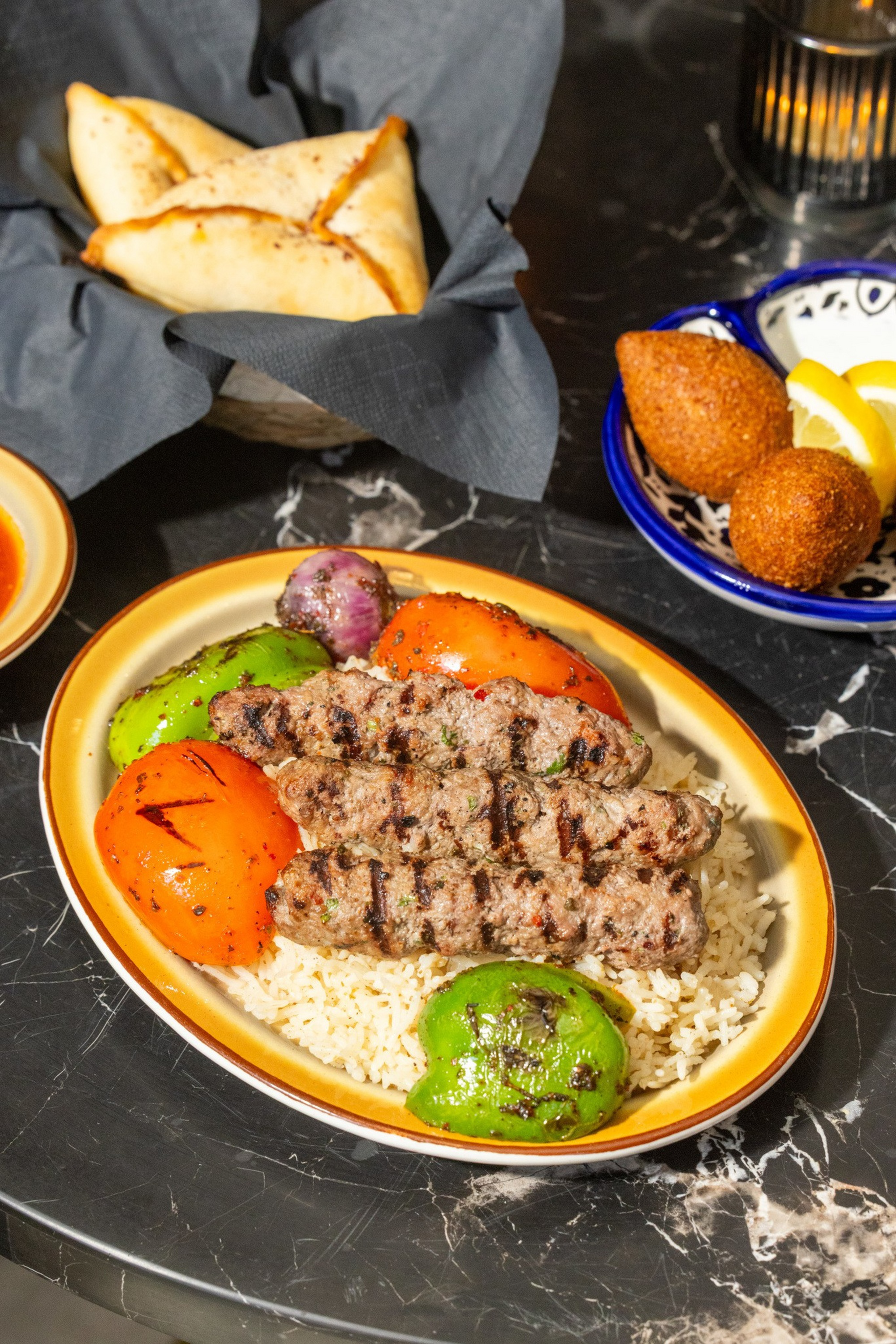 Ψητά κεμπάπ σερβιρισμένα με ρύζι με απανθρακωμένες πράσινες πιπεριές, ντομάτες πορτοκαλιού και ένα μικρό ψητό κρεμμύδι, συνοδευόμενα από γεμιστά γλυκά και τηγανητές κροκέτες με φέτες λεμονιού.