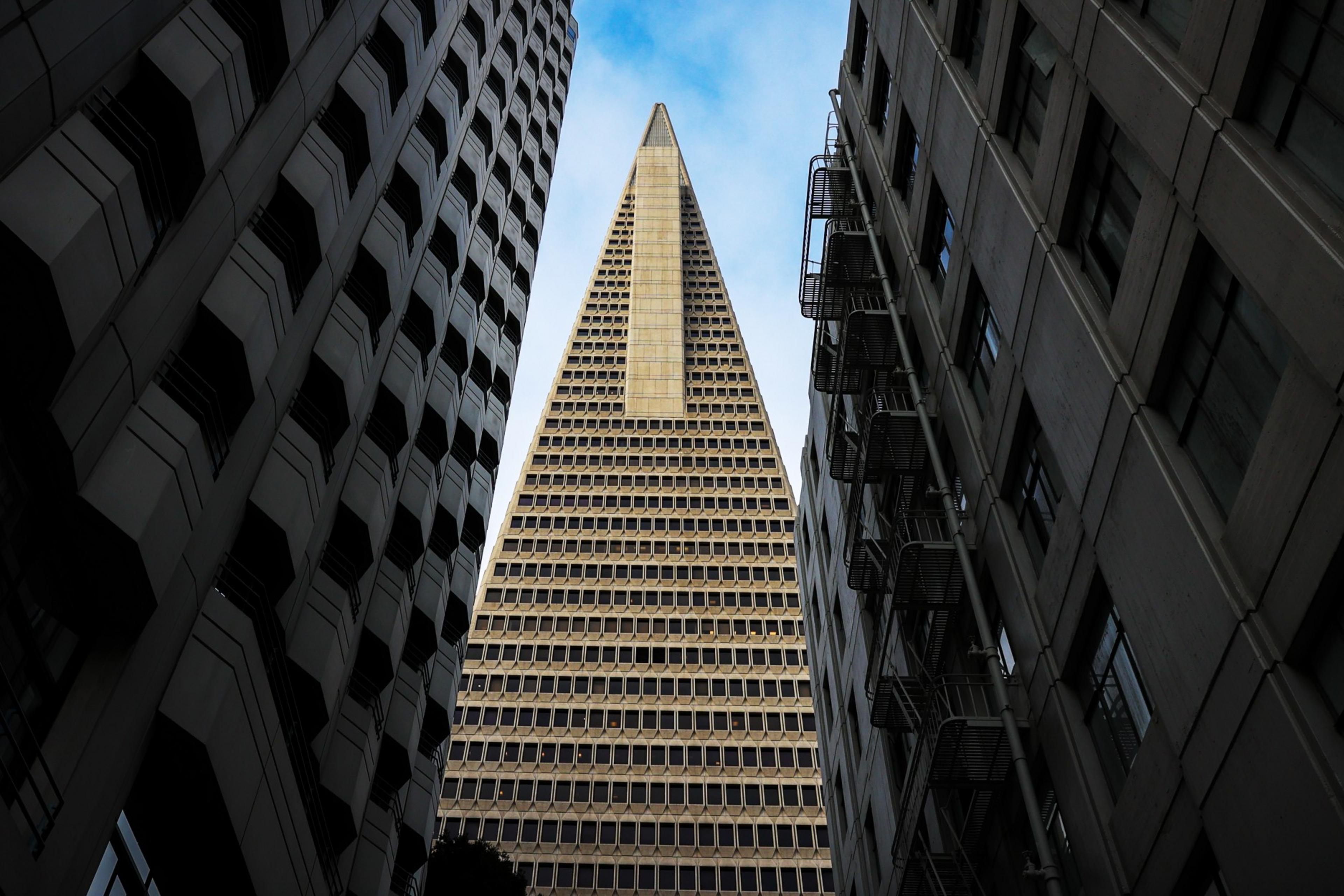 A towering skyscraper flanked by two buildings under a blue sky, viewed from a low angle.