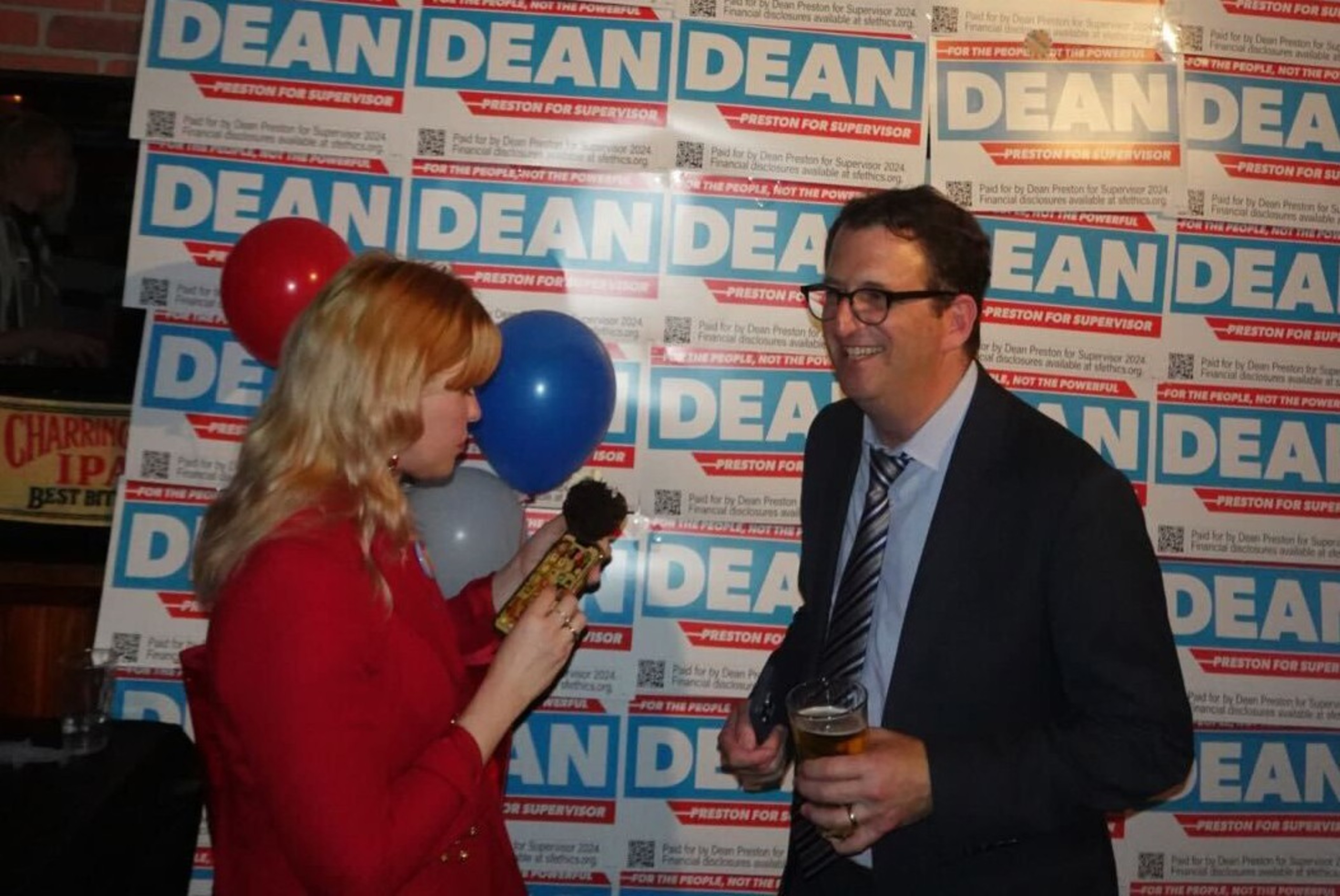 A woman in red holds a microphone, interviewing a man in glasses with a drink. They are in front of a &quot;Dean for Supervisor&quot; campaign sign.