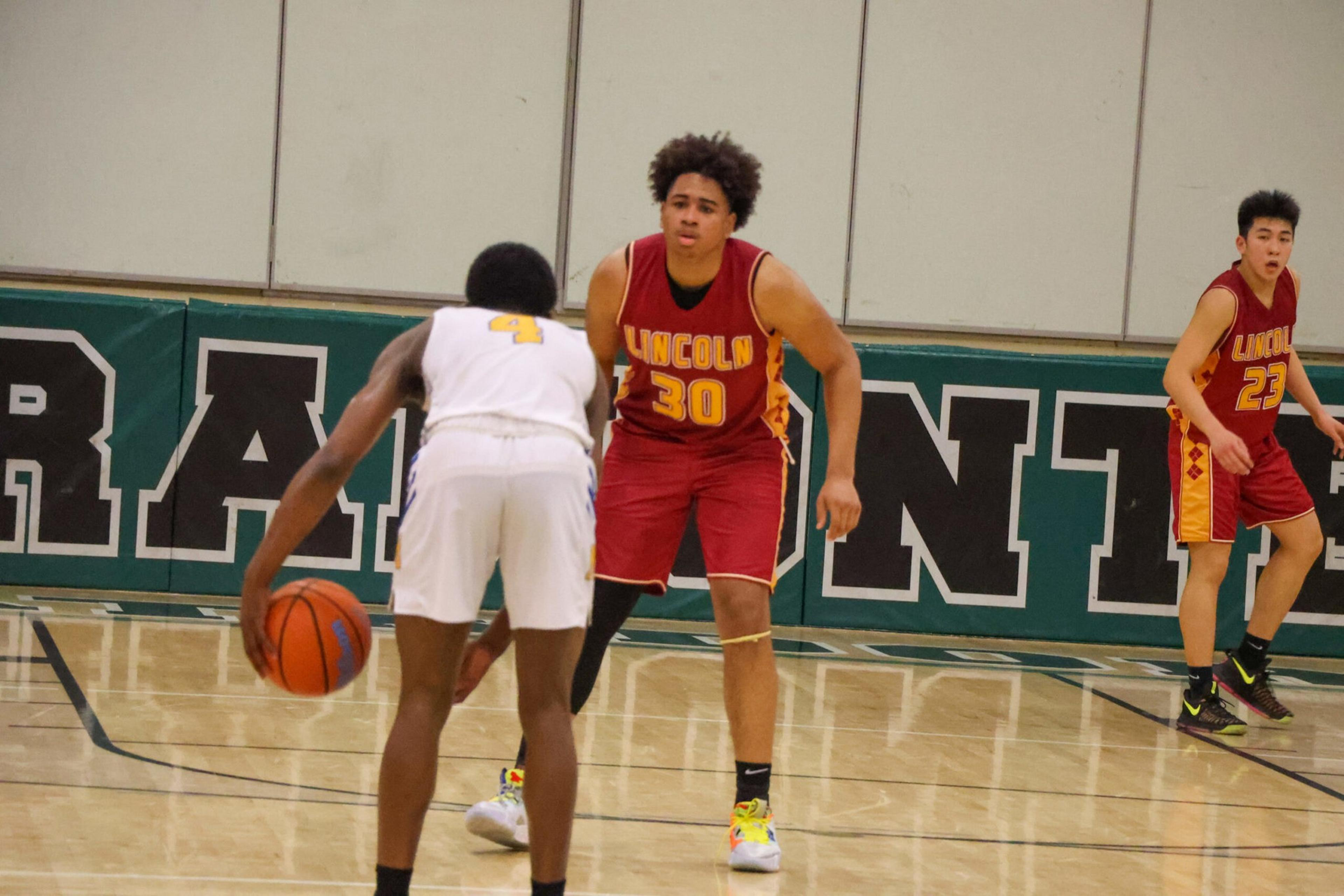A basketball player in a white uniform dribbles the ball while being closely guarded by a player in a red "Lincoln" jersey, number 30, with another teammate nearby.