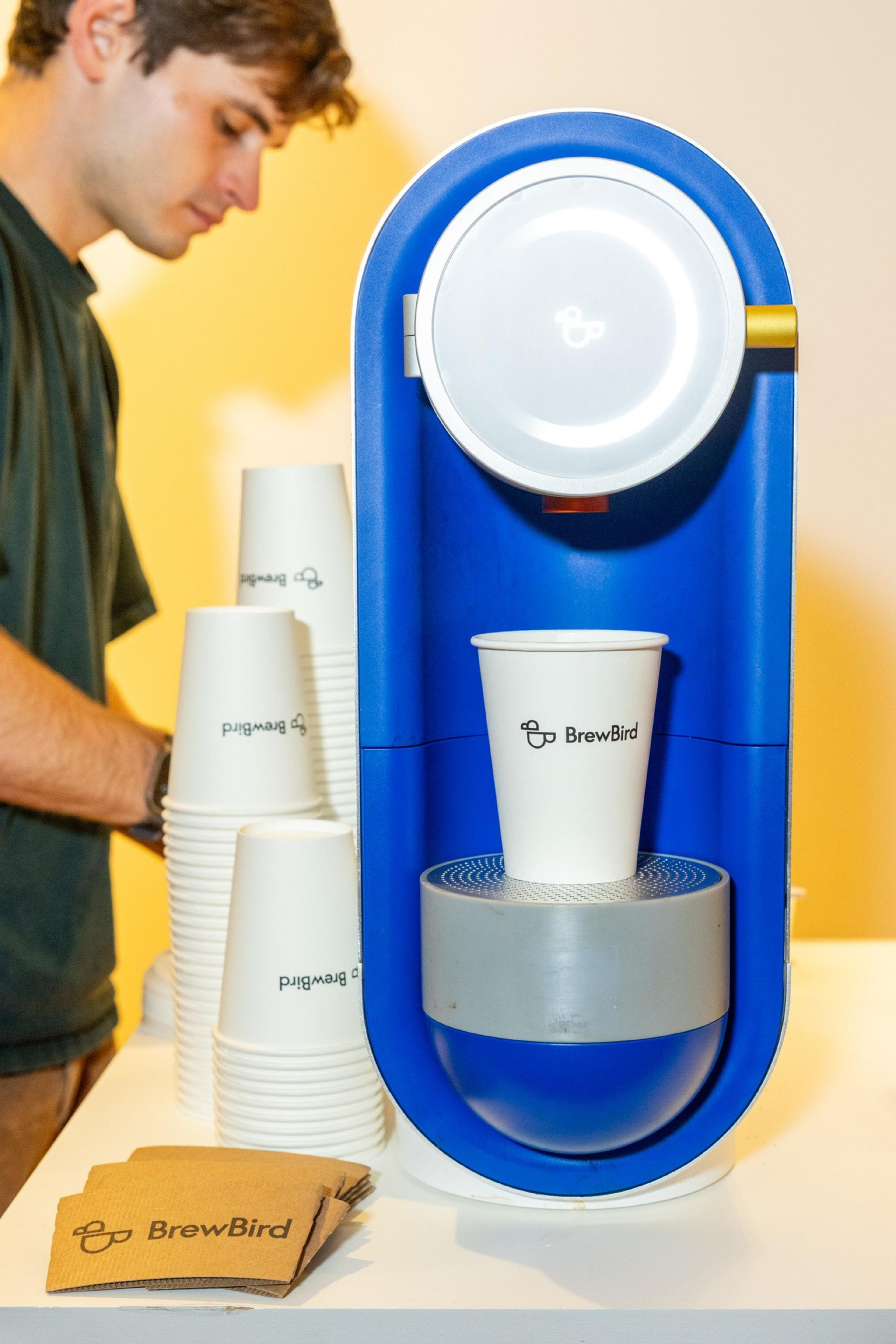 A blue and white BrewBird coffee machine dispenses coffee into a branded paper cup, with more cups and napkins stacked beside it and a person in the background.