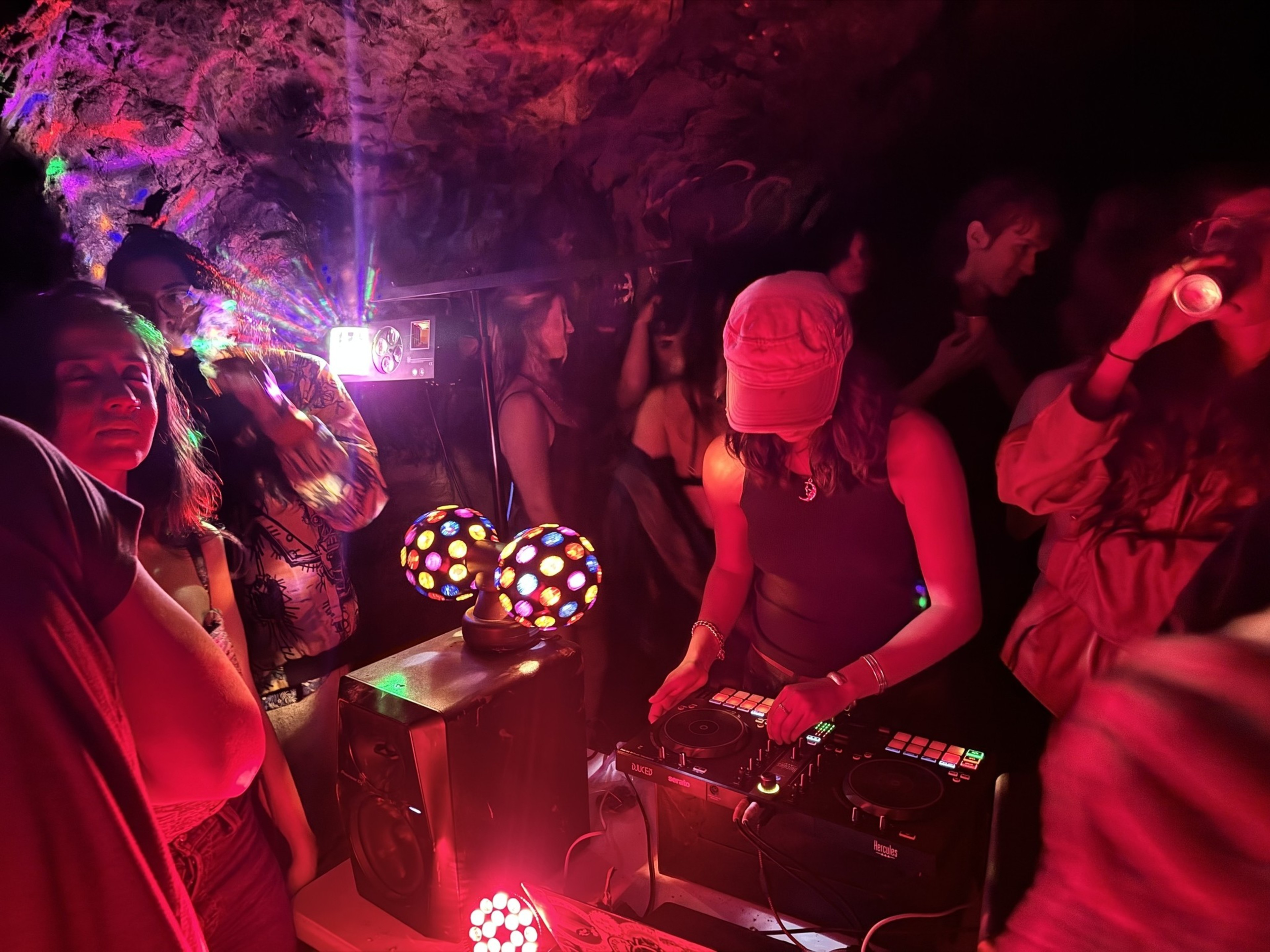 People gathered in a dimly lit, colorful underground party, with vibrant lights, a DJ wearing a cap playing music, and attendees dancing and socializing around.