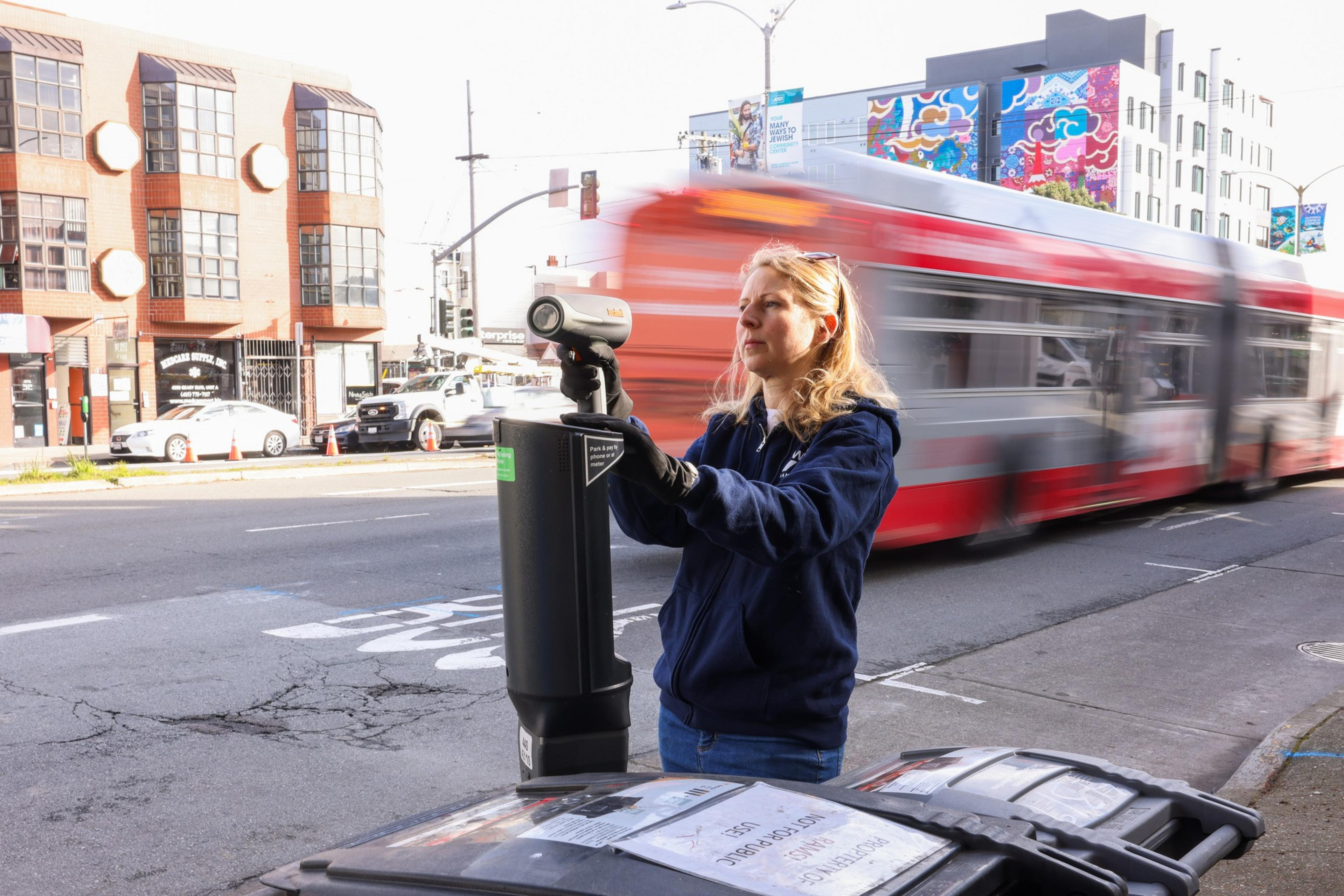 A woman in a navy jacket holds a small device on a street with a blurred red bus passing by. Buildings and cars are visible in the background.