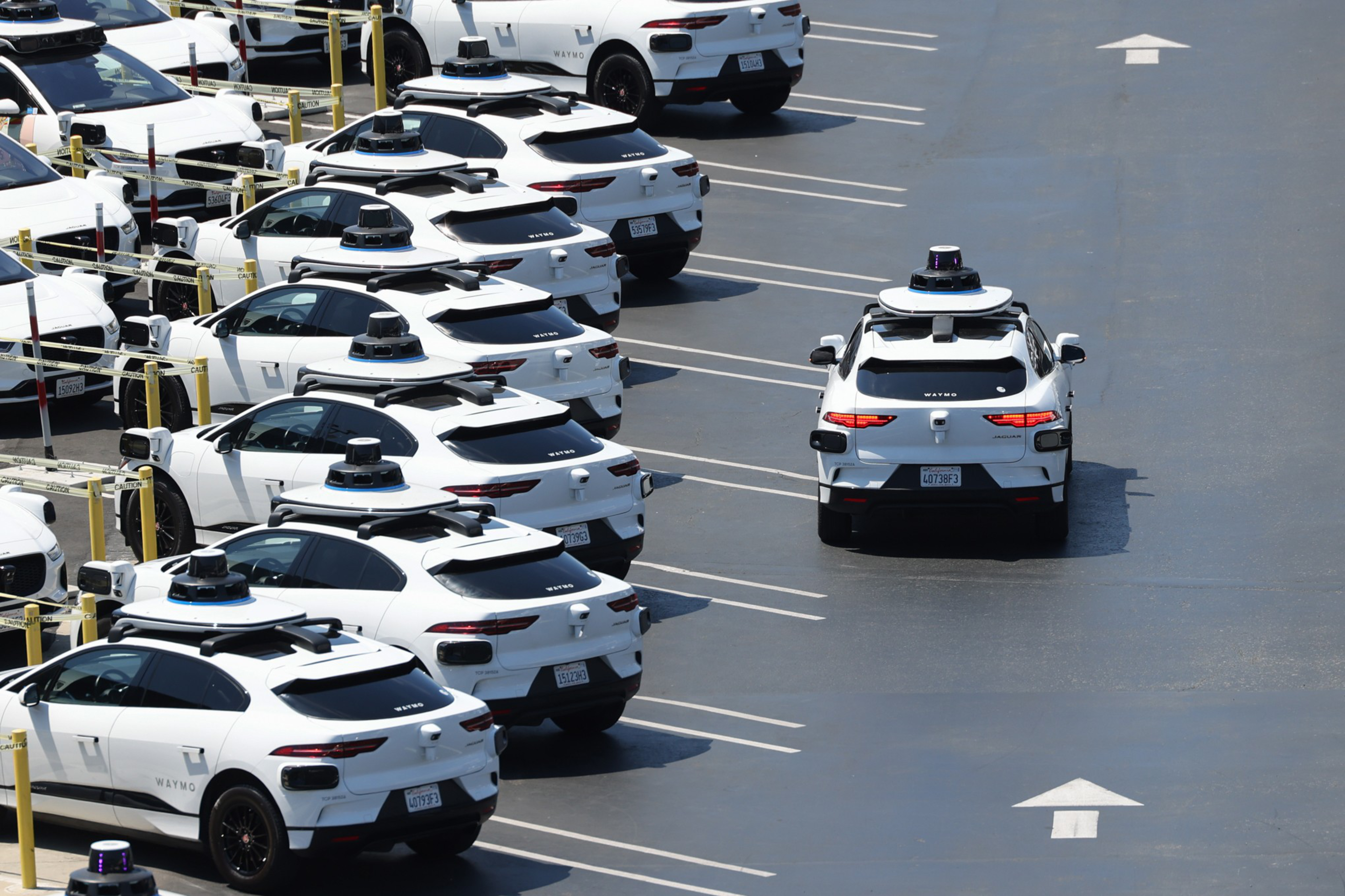Multiple white self-driving cars with roof sensors are parked in a lot, while one similar car drives forward in a marked lane.
