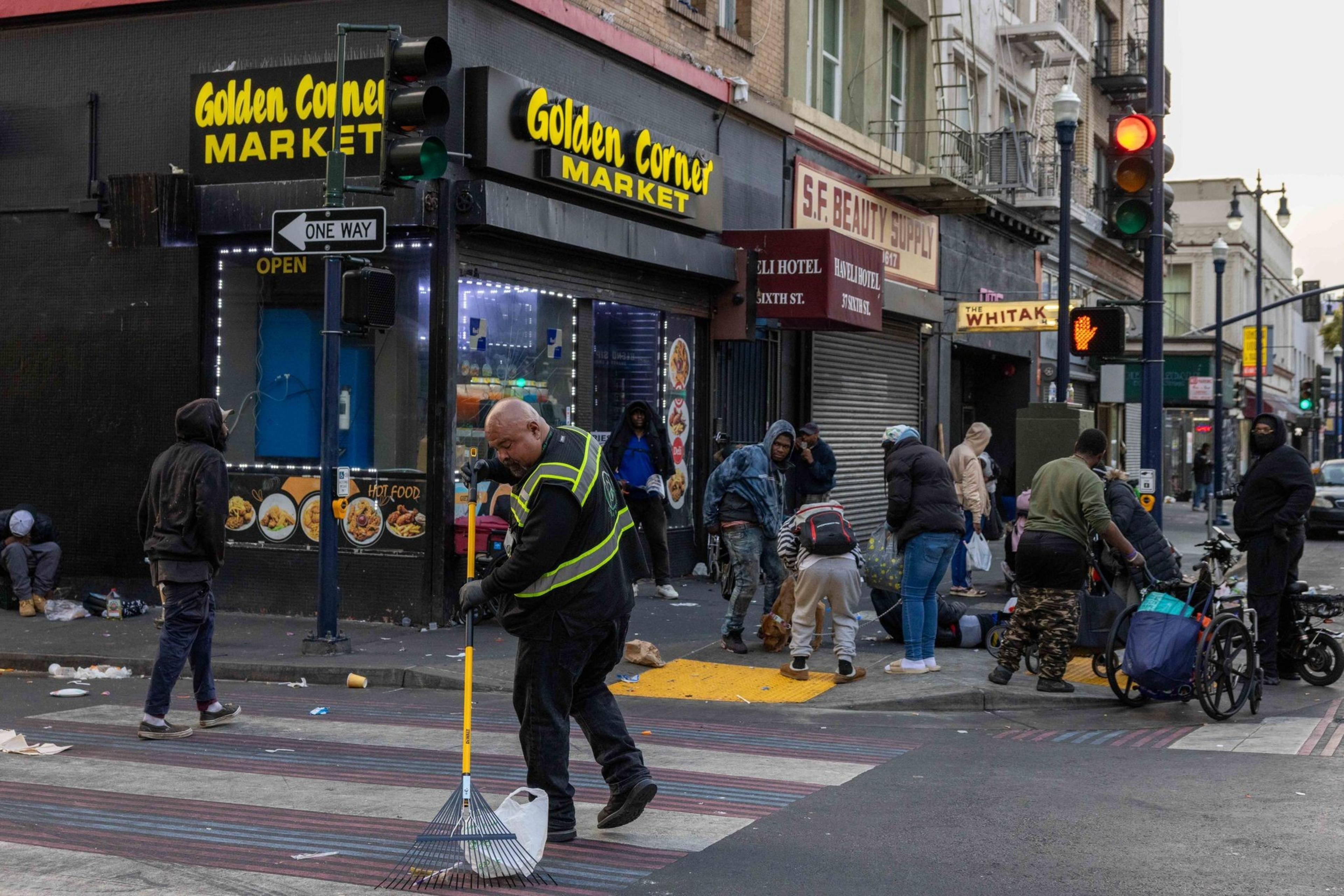 A man sweeps a street in front of &quot;Golden Corner Market&quot; while people gather nearby, some sitting, standing, or crouching, amidst scattered litter.