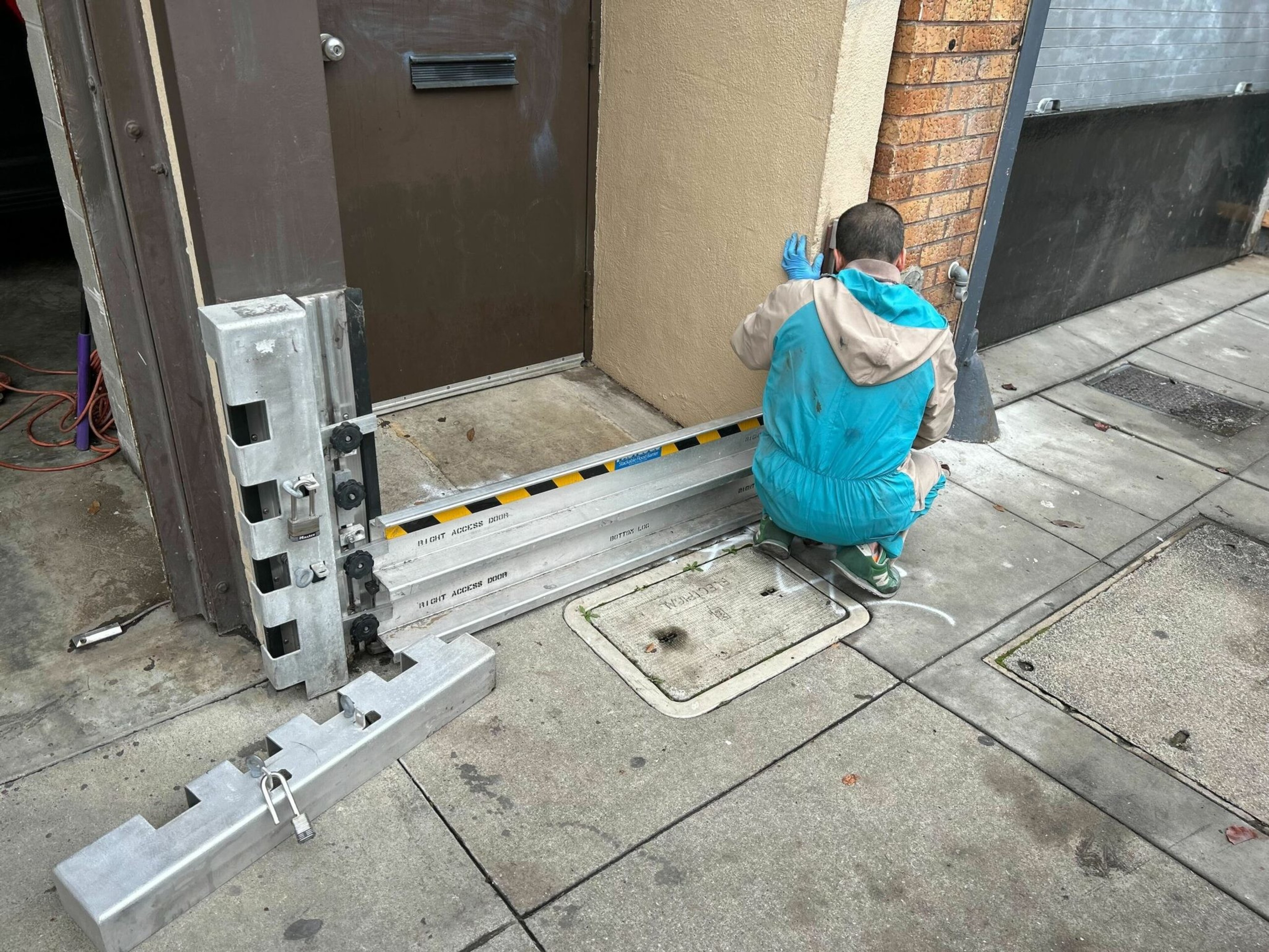 A person in a blue and beige jacket is crouched by a brick wall, installing or adjusting metal flood barriers on a sidewalk near a door.