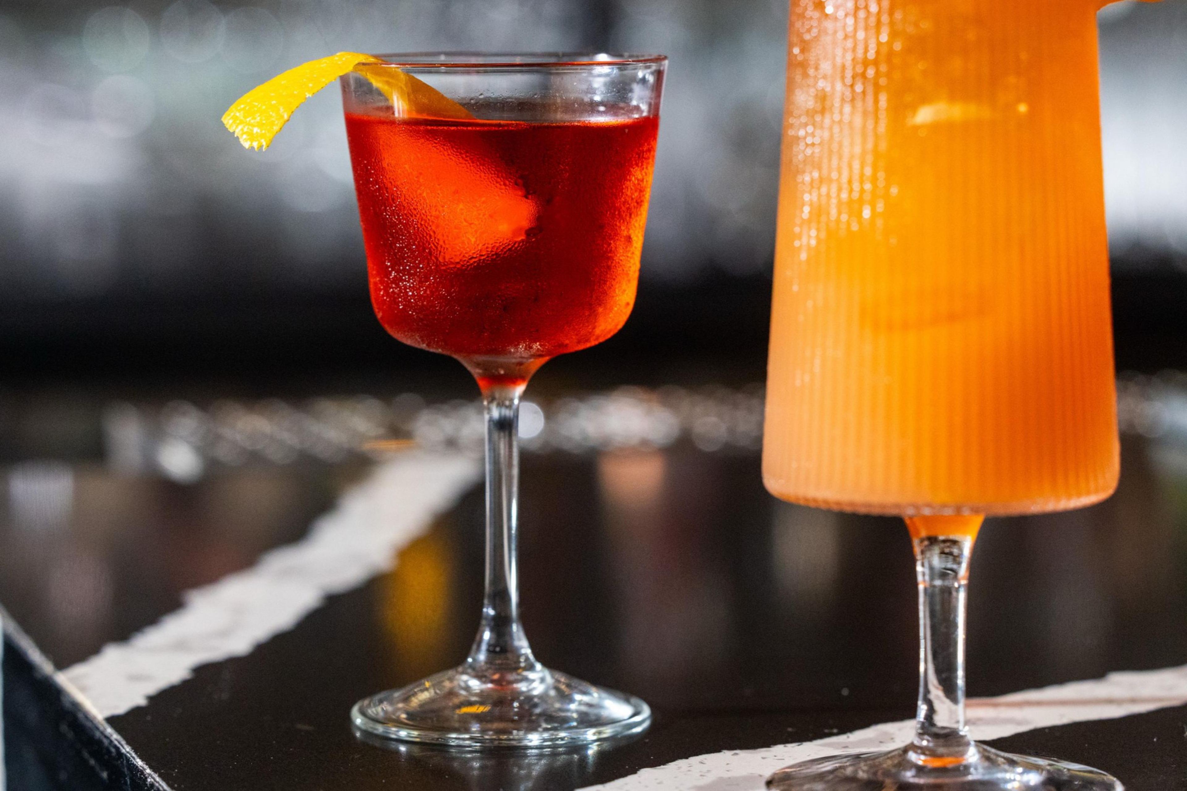 A red cocktail in a coupe glass with an orange peel garnish sits next to a tall orange drink with ice in a ribbed glass on a dark bar counter.