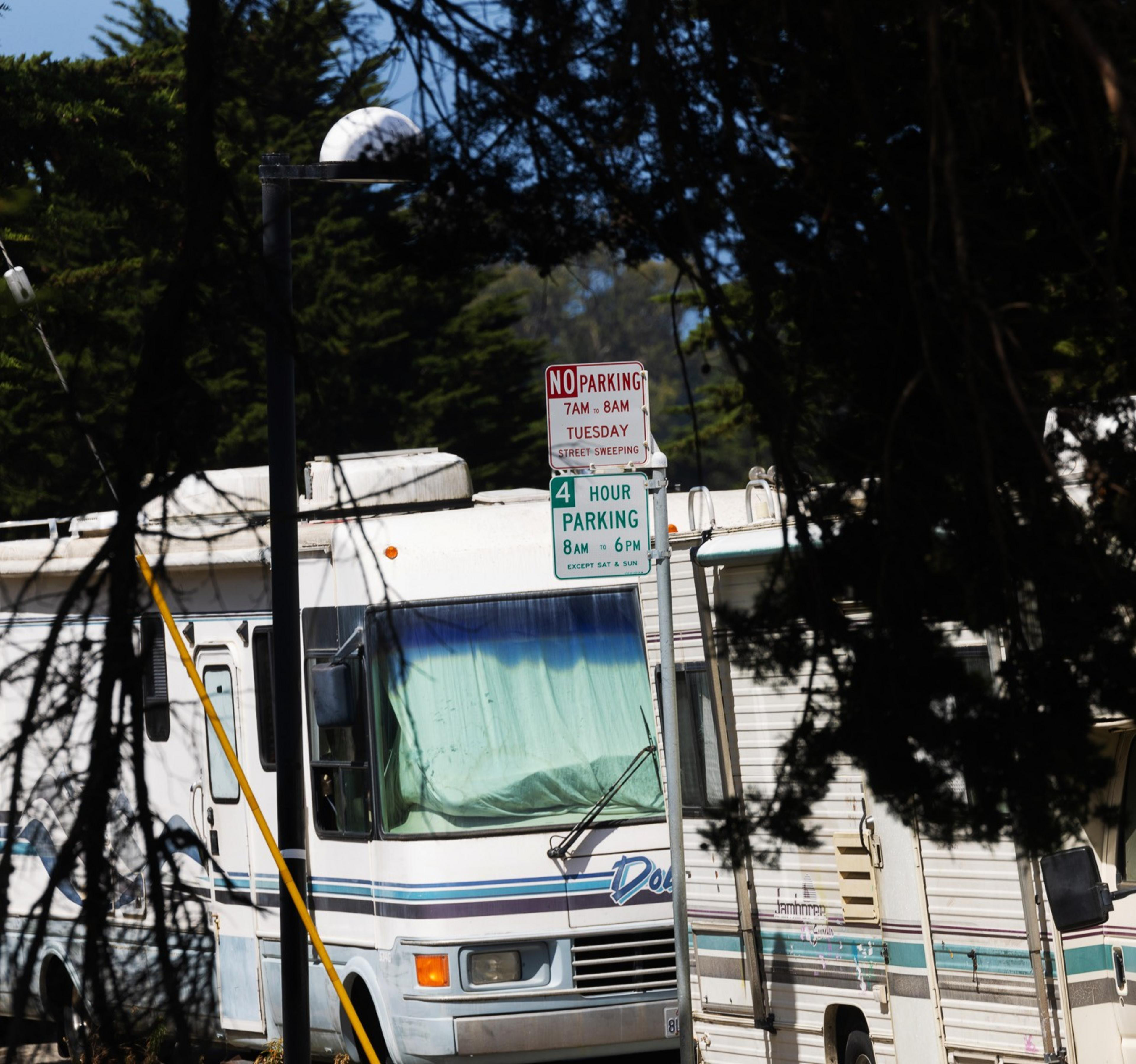The image shows an RV parked on the street, partially obscured by trees. There are parking restriction signs indicating no parking on Tuesdays from 7-8 AM and a four-hour limit from 8 AM to 6 PM.