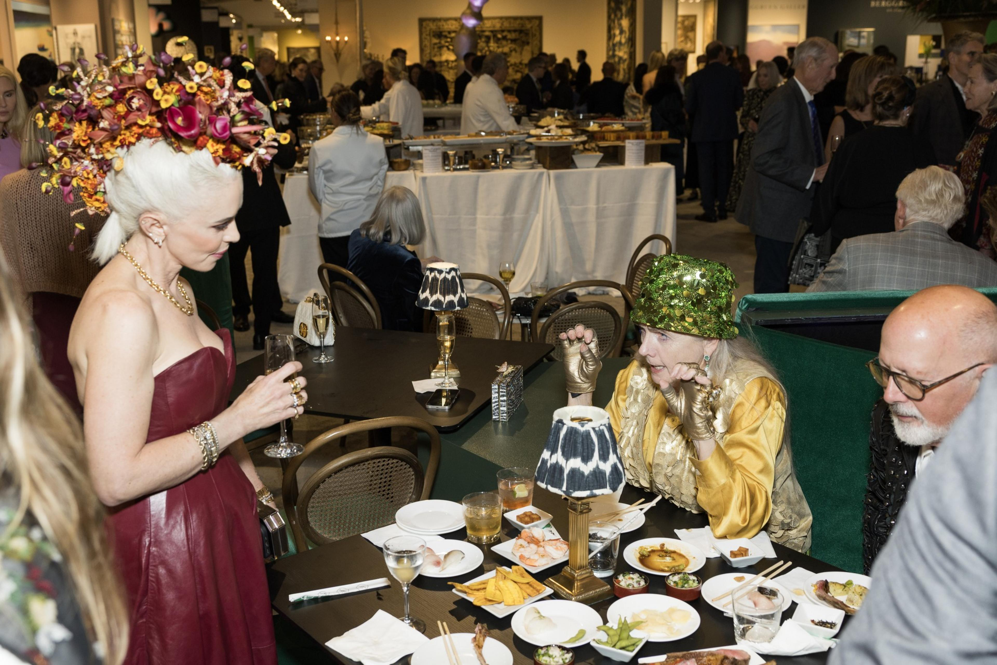 A woman in a red dress and floral headpiece holds a drink, while others at a table with appetizers and drinks chat in a dimly lit, busy event space.