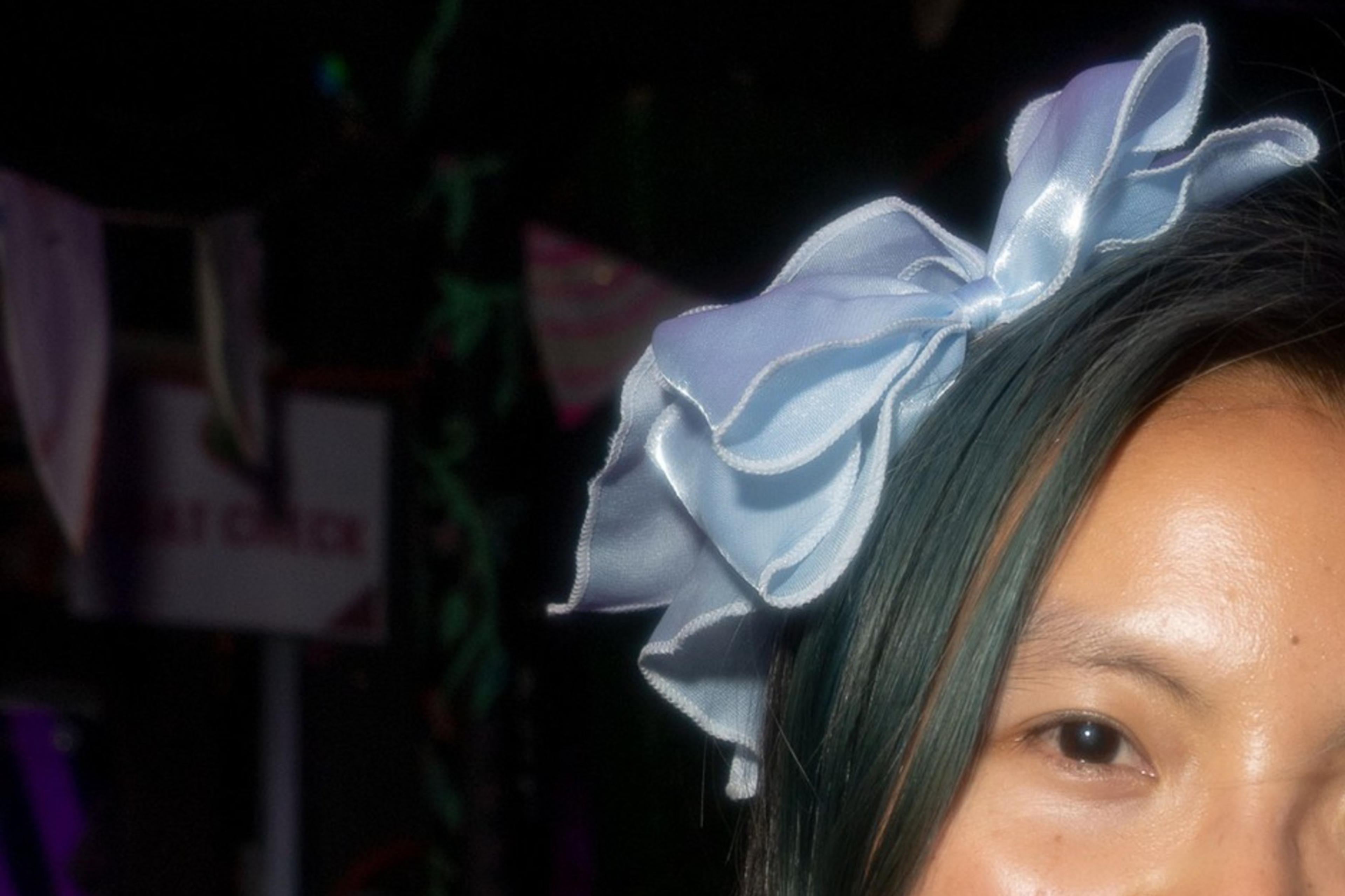 A woman with long hair holds a blue lollipop in her mouth. She wears a large bow and a smartwatch. A neon sign with "Fuzzy Plac." is visible behind her.