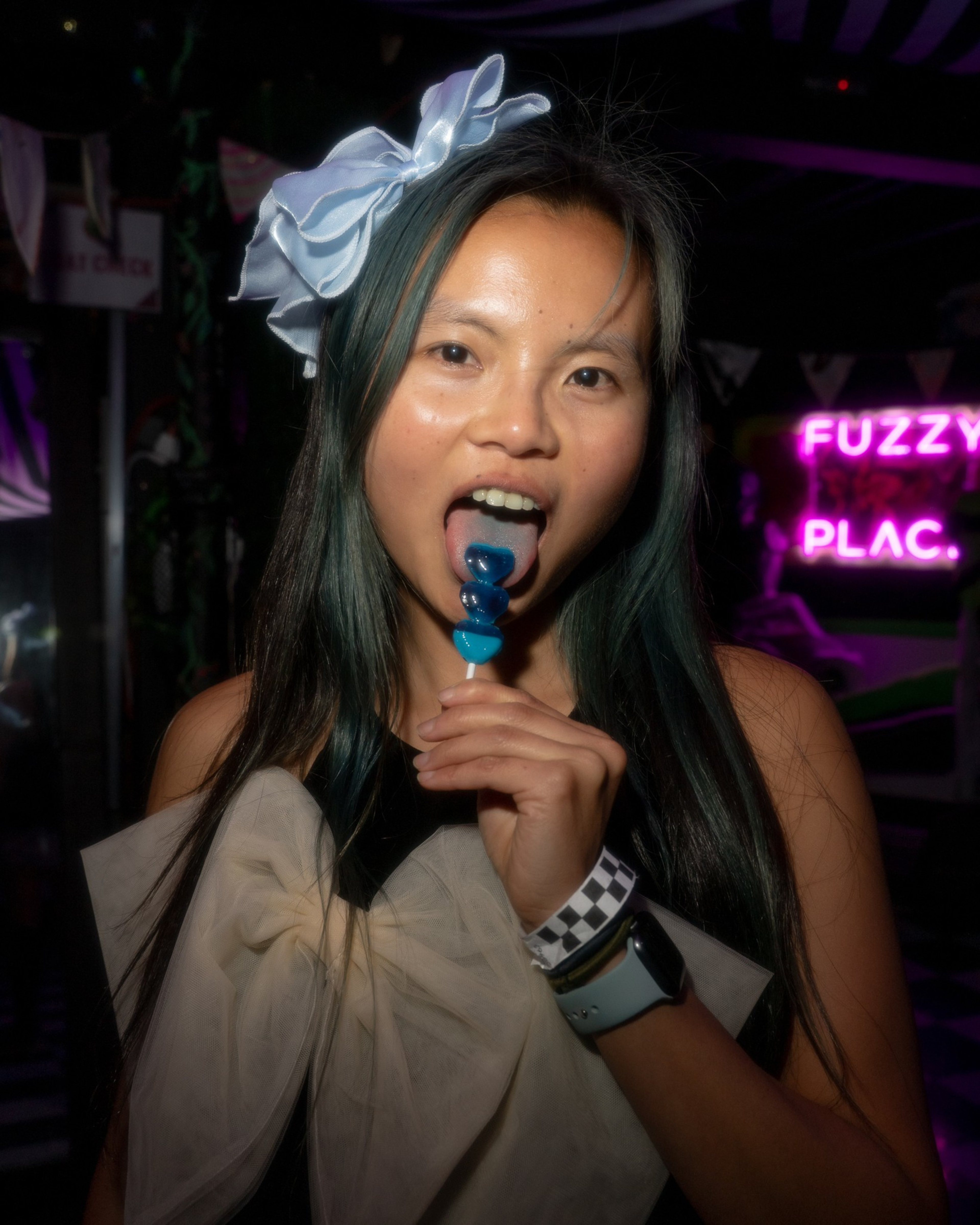 A woman with long hair holds a blue lollipop in her mouth. She wears a large bow and a smartwatch. A neon sign with "Fuzzy Plac." is visible behind her.