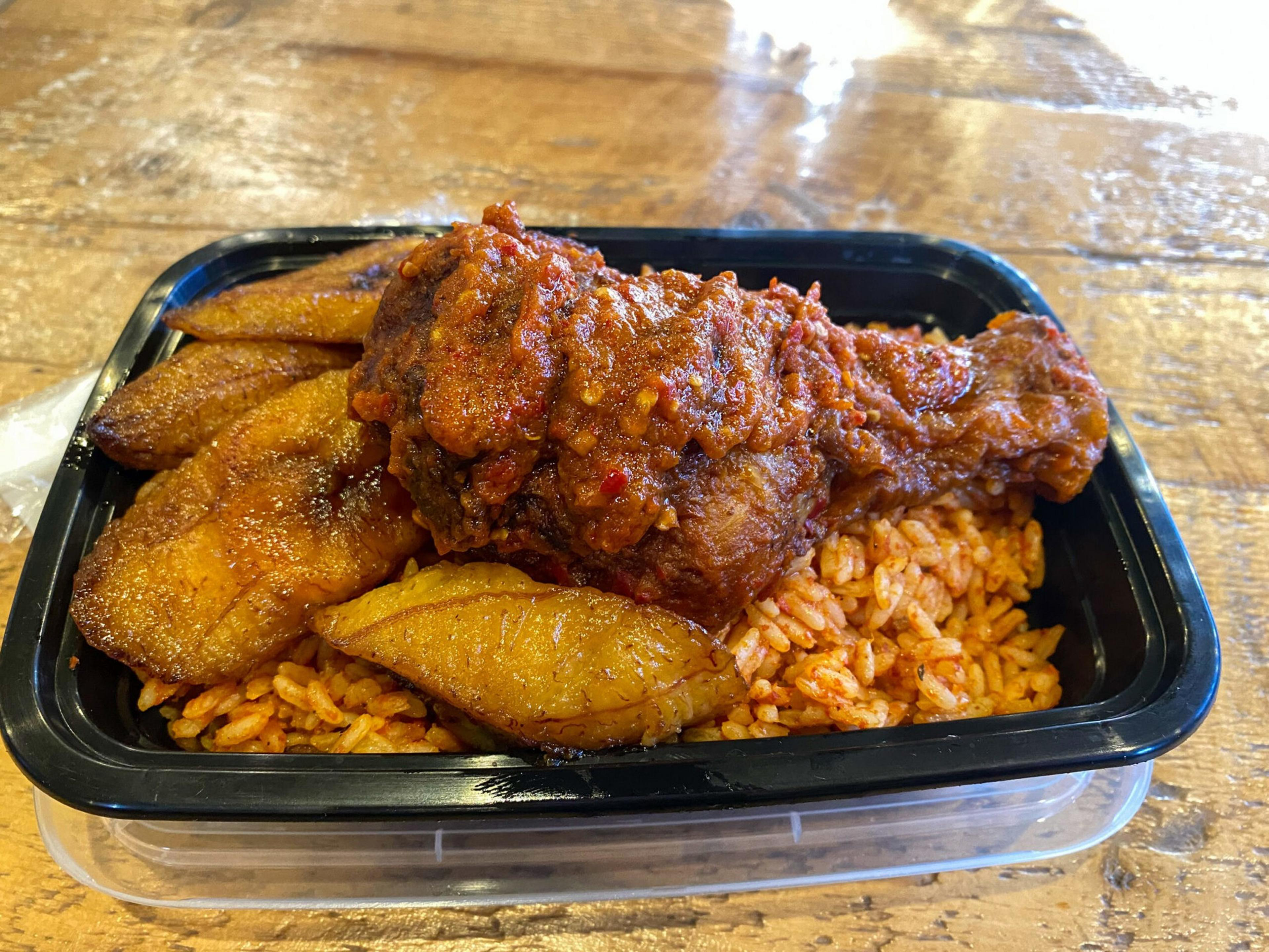 Jollof rice topped with a spicy fried chicken drumstick and several pieces of fried plantain in a black plastic container.