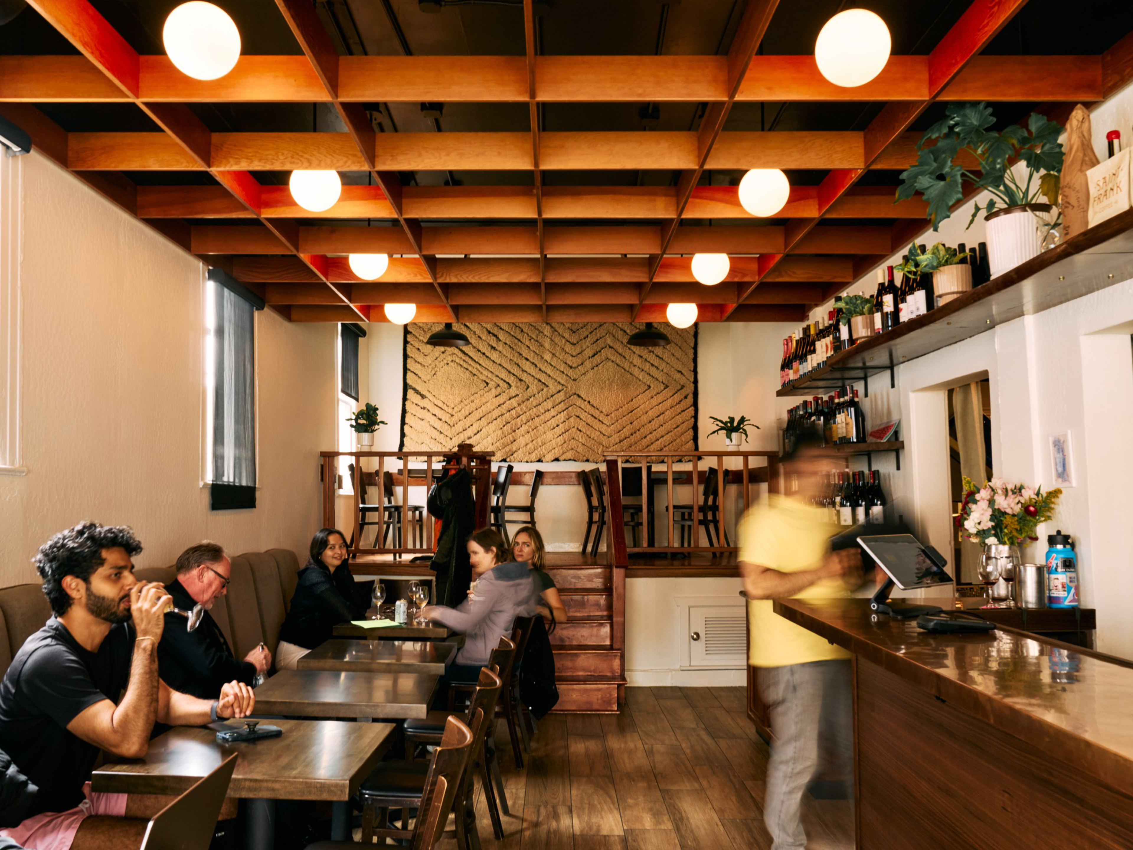 A cozy café with wooden tables and chairs, warm lighting, plants on shelves, and people sitting and chatting while a blurry server stands at the counter.