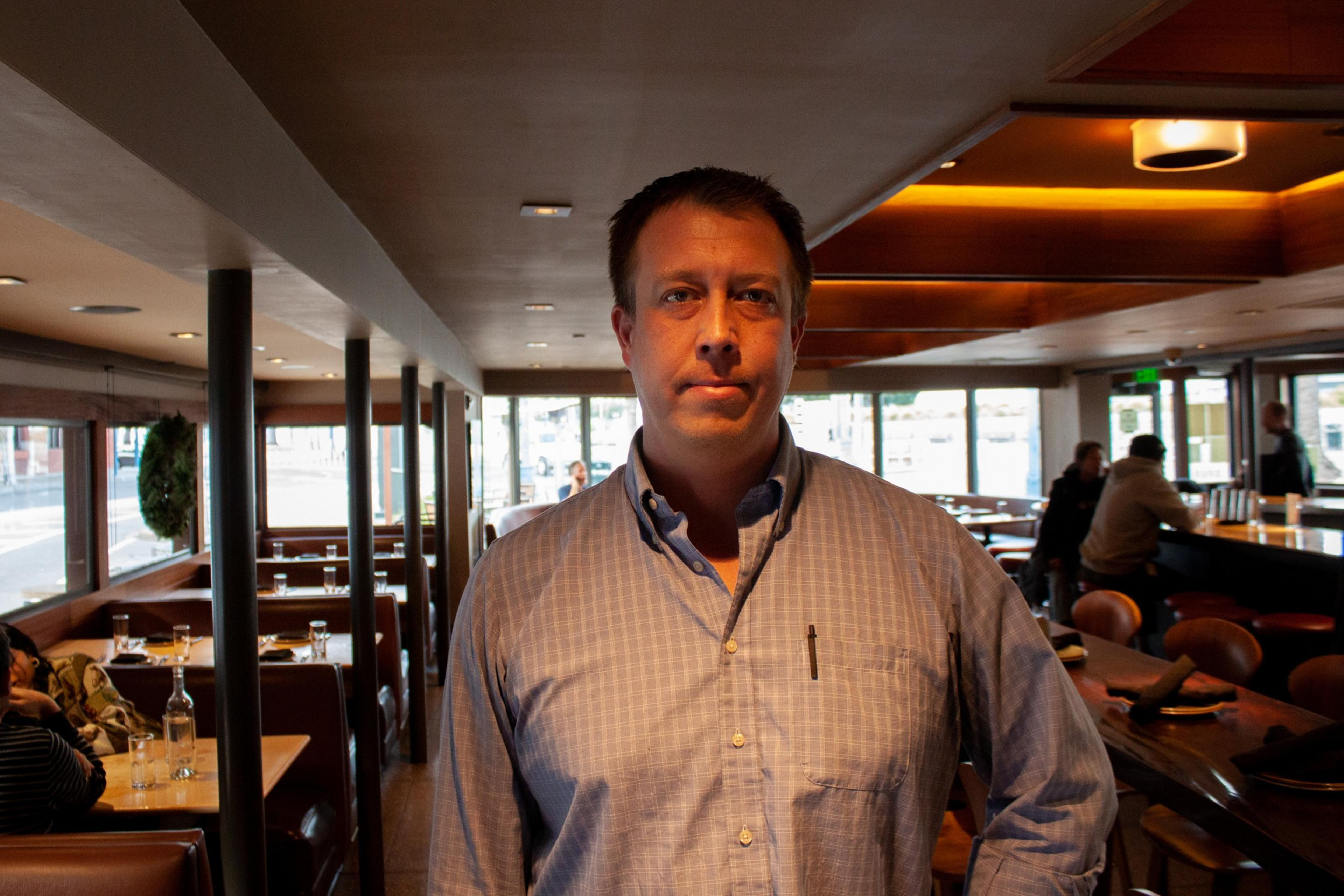 A man in a buttoned-up, light blue checkered shirt stands inside a warmly lit restaurant with wooden tables and large windows.