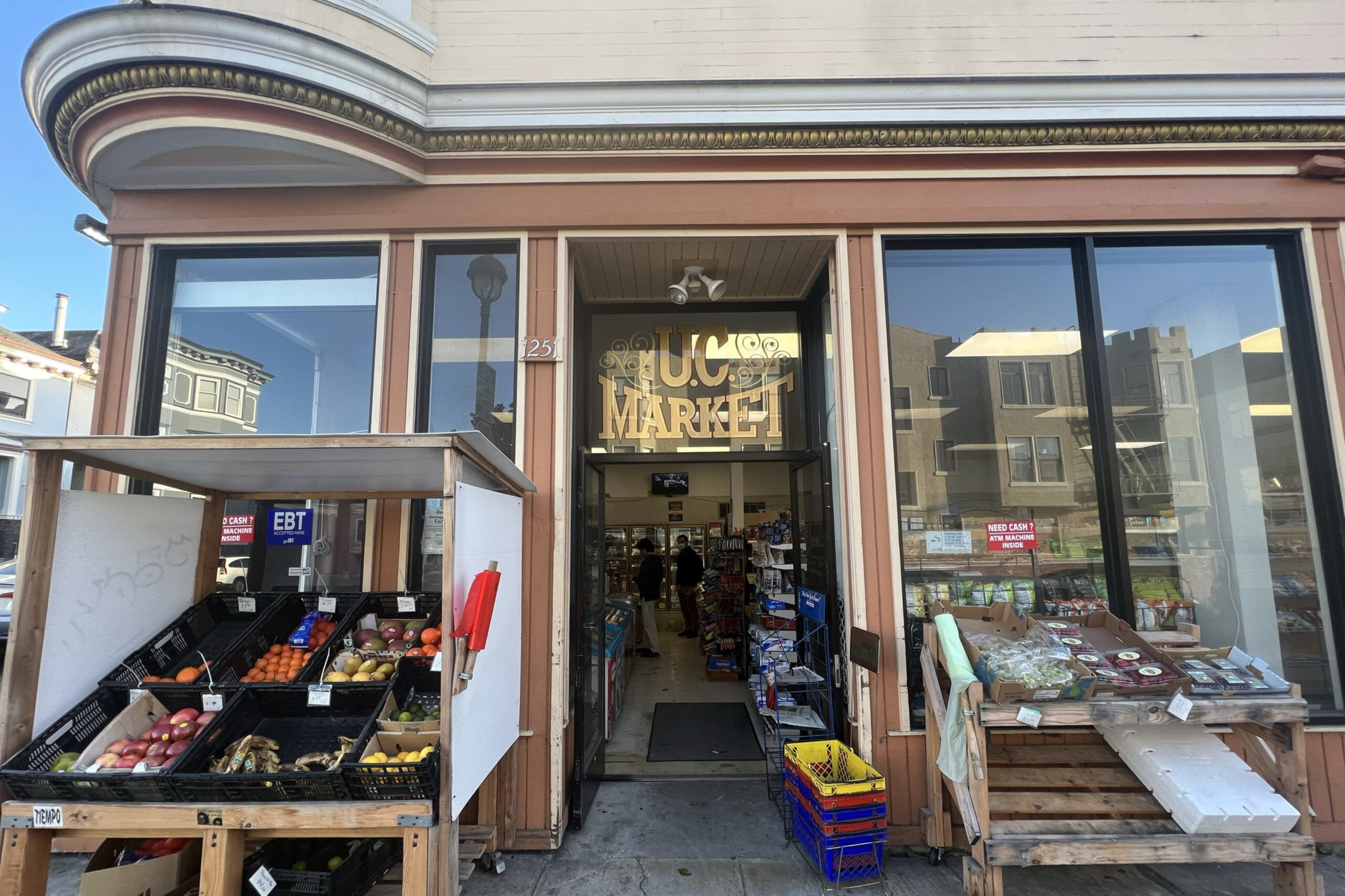 A storefront labeled "U.C. Market" displays fresh fruits and vegetables on wooden shelves outside, with an open door leading inside a small grocery store.