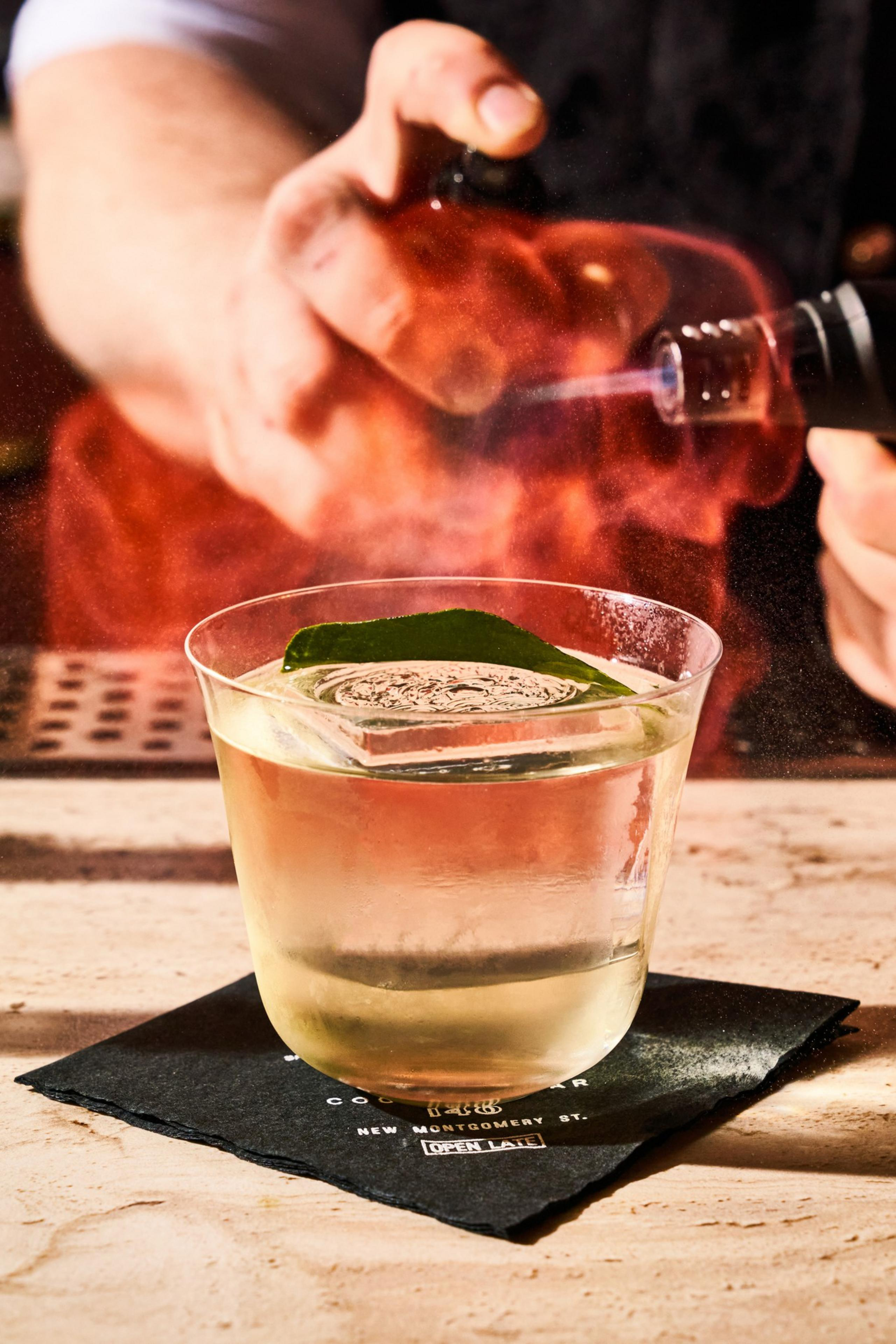 A cocktail in a clear glass with a large ice cube and a green leaf on top sits on a black napkin. A hand is using a torch to create a smoky effect.
