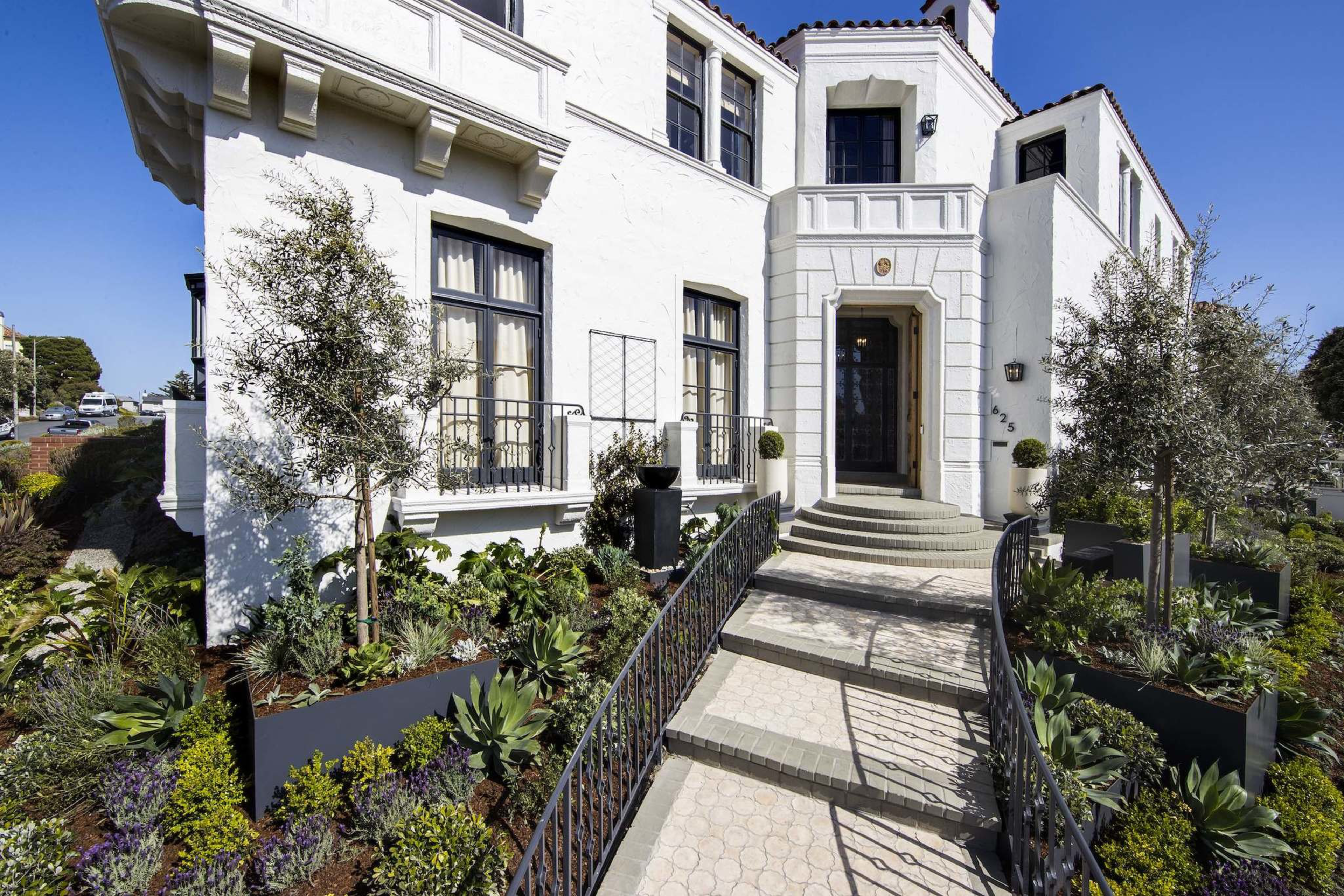 A white two-story house with black-trimmed windows, a curved walkway and steps, and neatly landscaped plants and trees on both sides.
