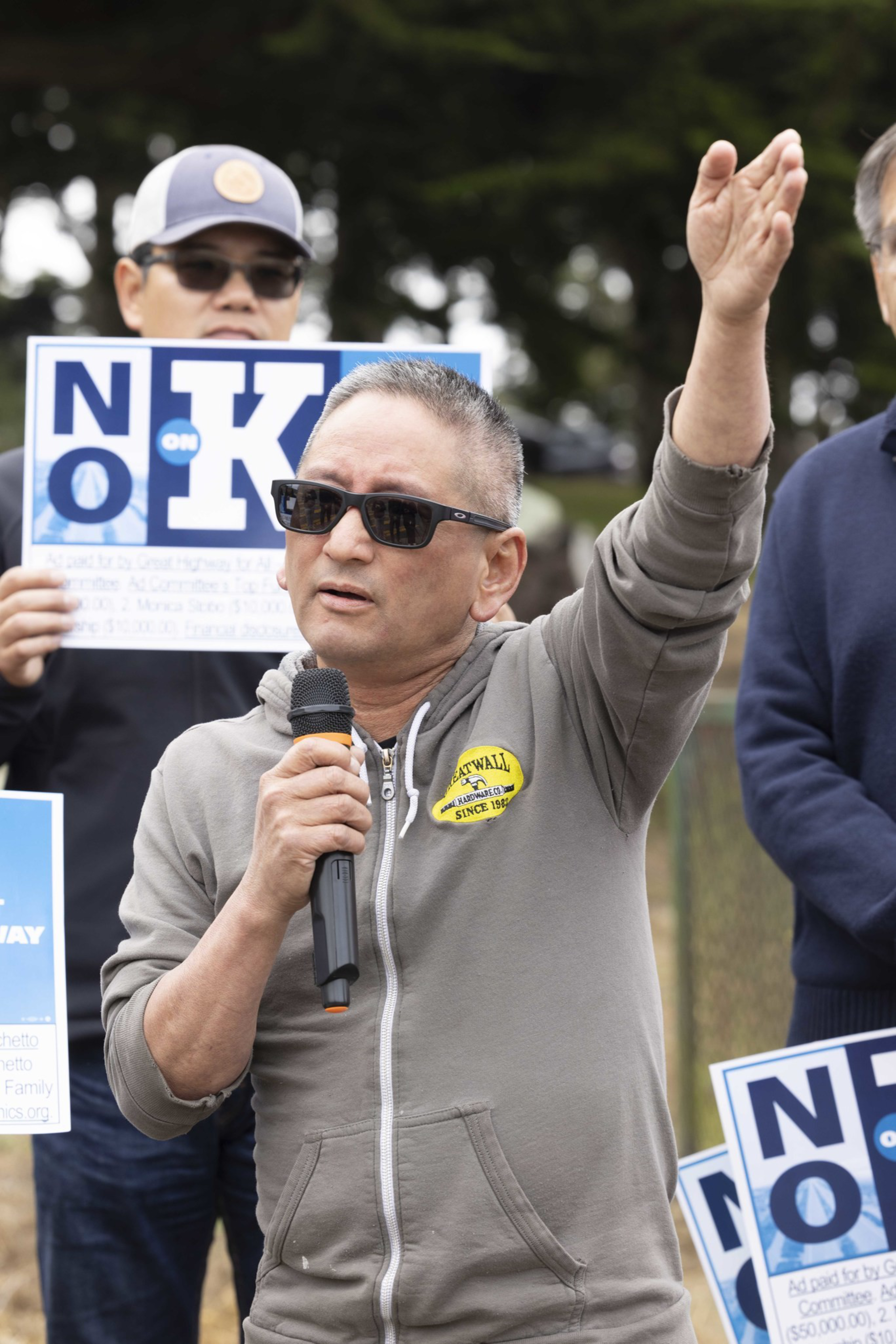 A man in a gray hoodie, speaking into a microphone with one hand raised, stands outdoors in front of a group. He is wearing sunglasses, and others hold &quot;No on K&quot; signs.