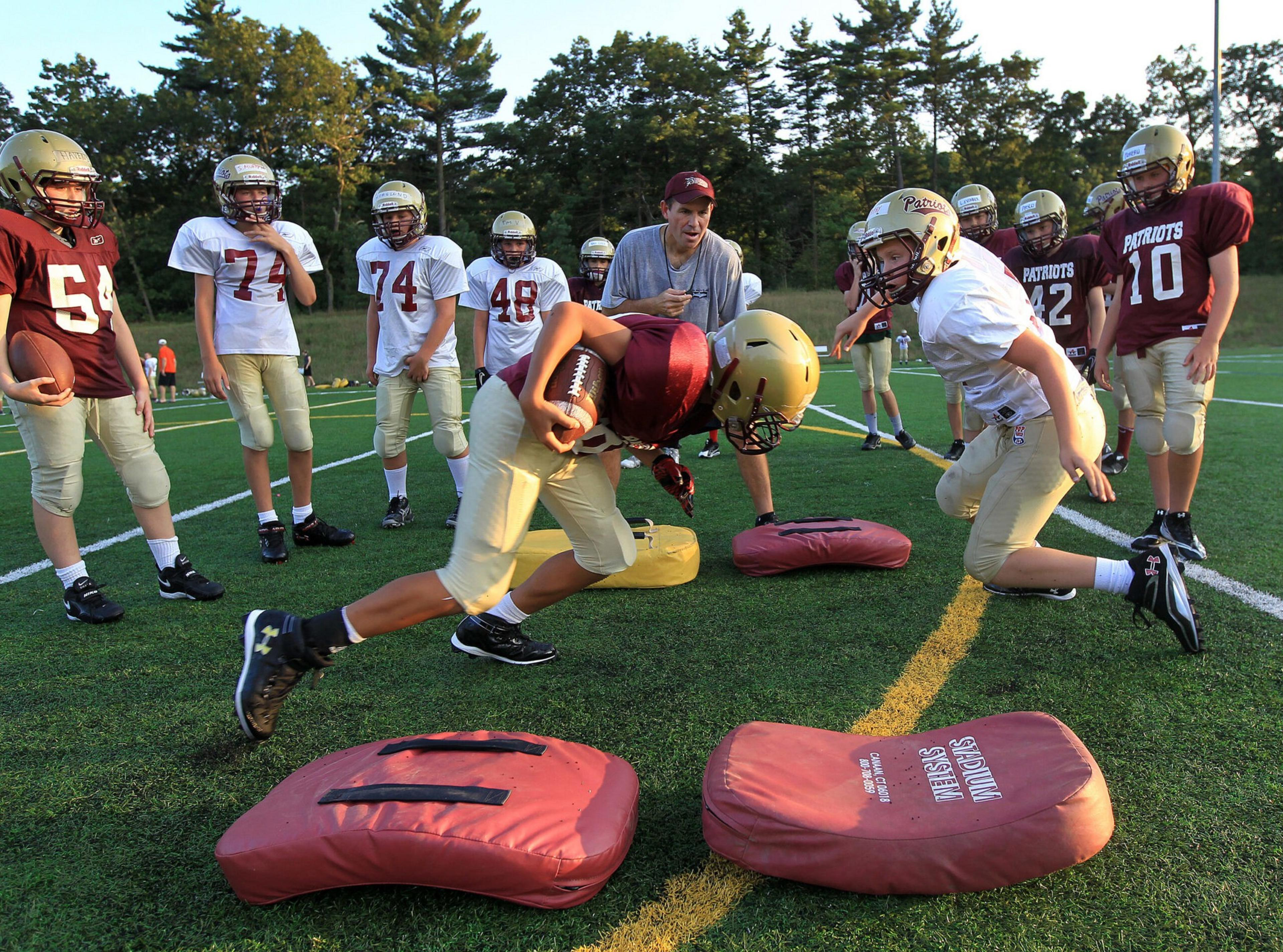 Young football players in helmets practice tackling drills on a field while a coach gives instructions and teammates watch.