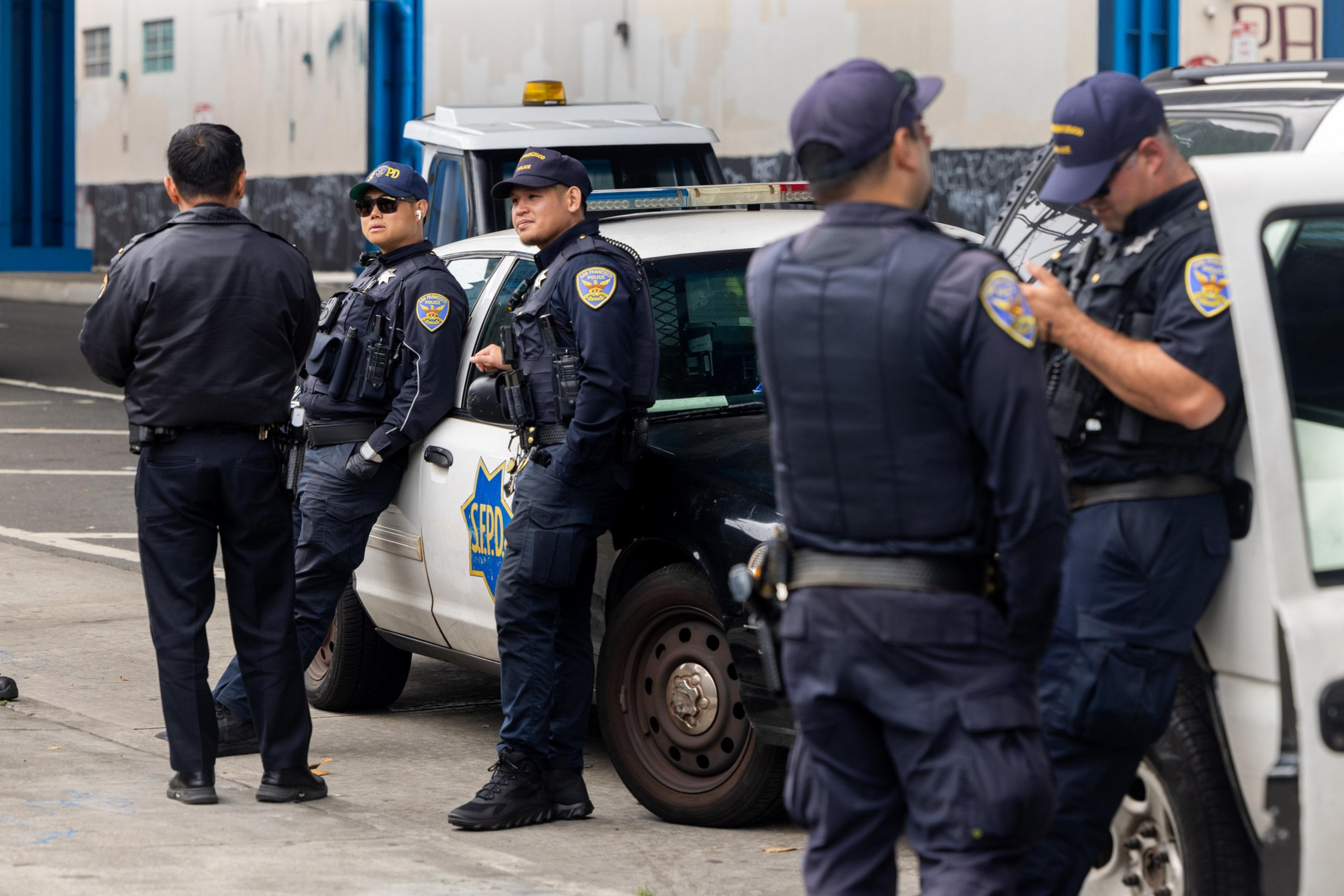 The image shows several police officers in uniform gathering around parked patrol cars in an urban setting, possibly engaged in a discussion or briefing.