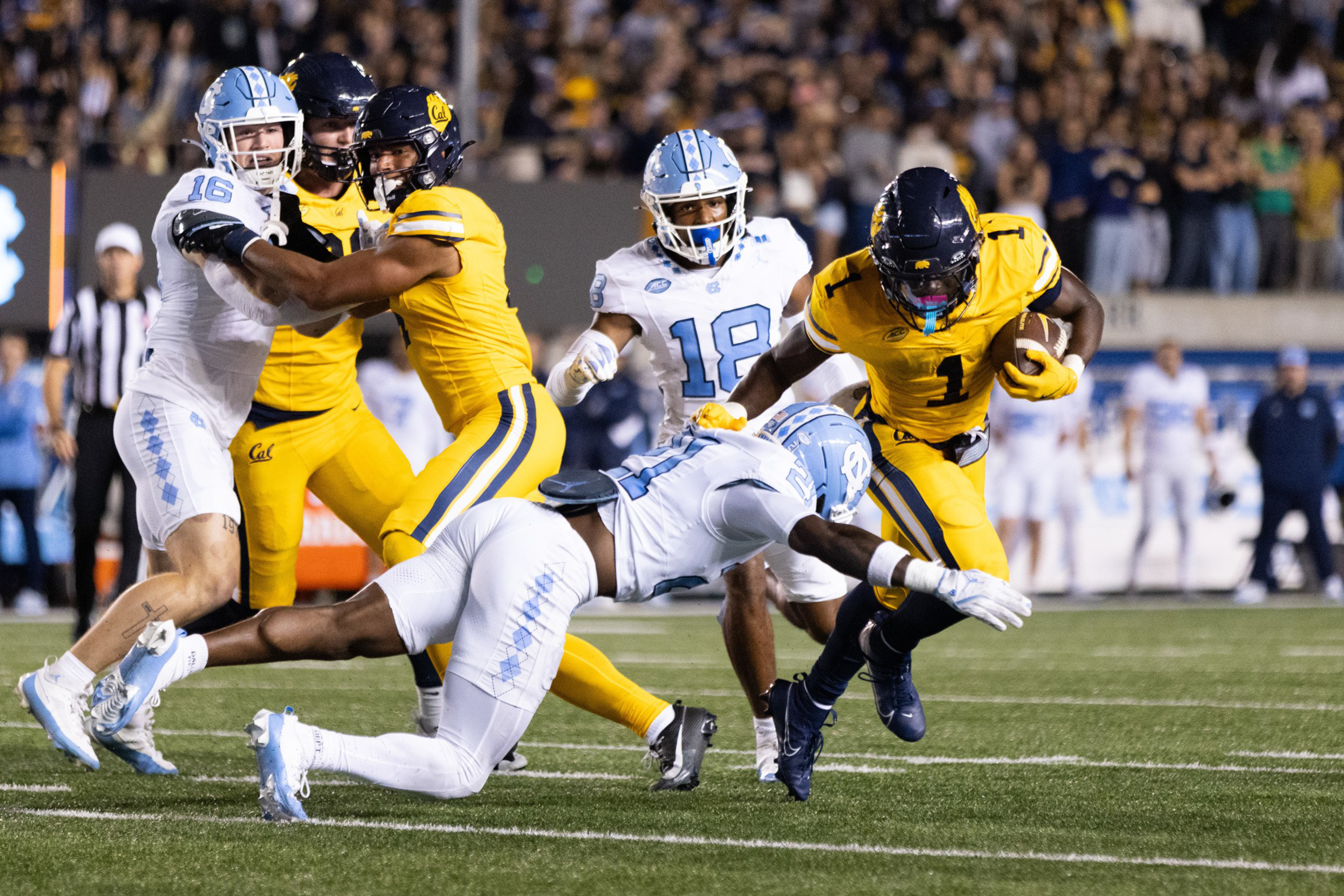 A player in yellow dodges a dive tackle from a player in white while two other players in yellow and white watch nearby on a football field.