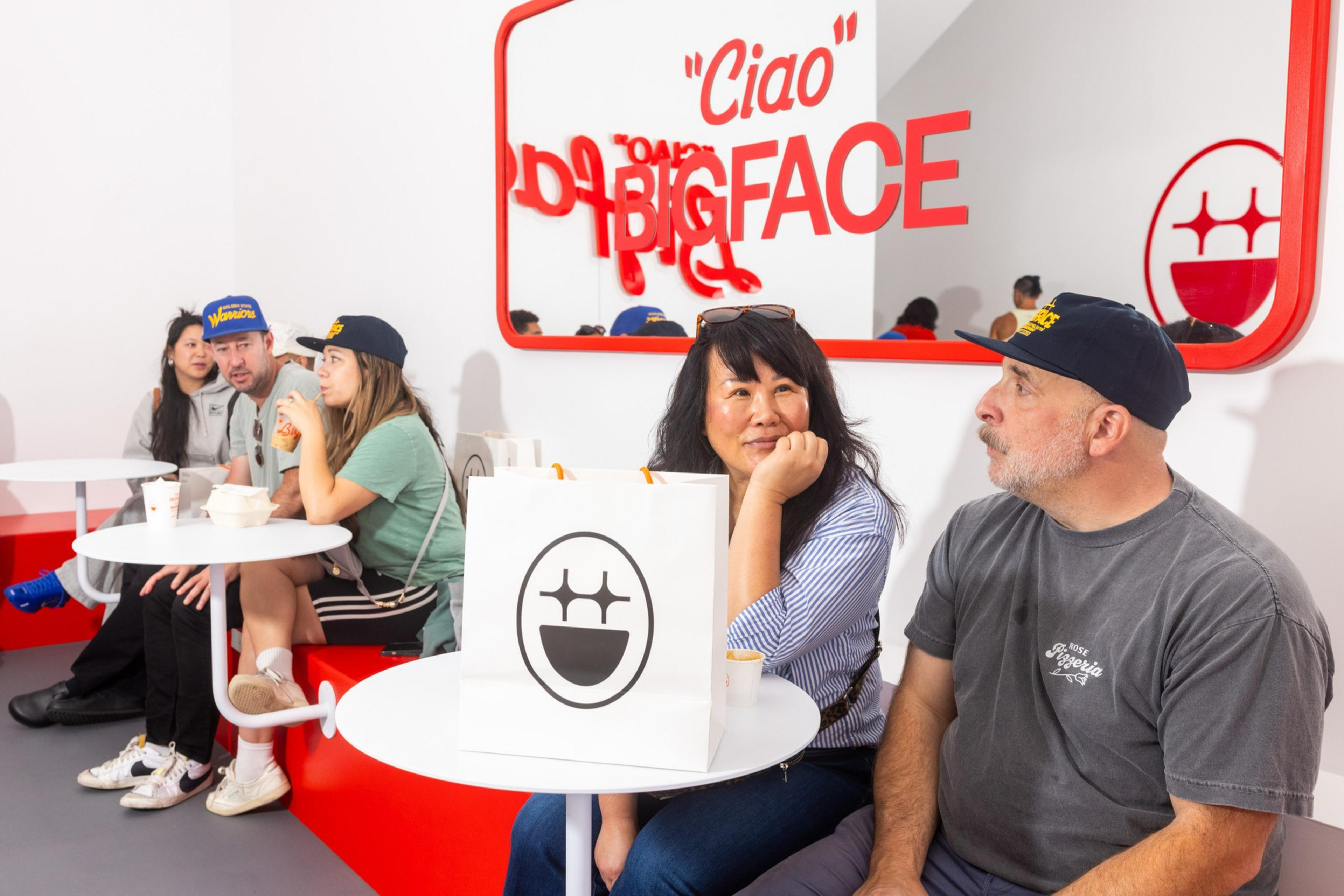 People sit at red and white tables, chatting and eating. A bag with a smiley logo and "Ciao BIGFACE" is on the wall. They wear casual clothes and caps.