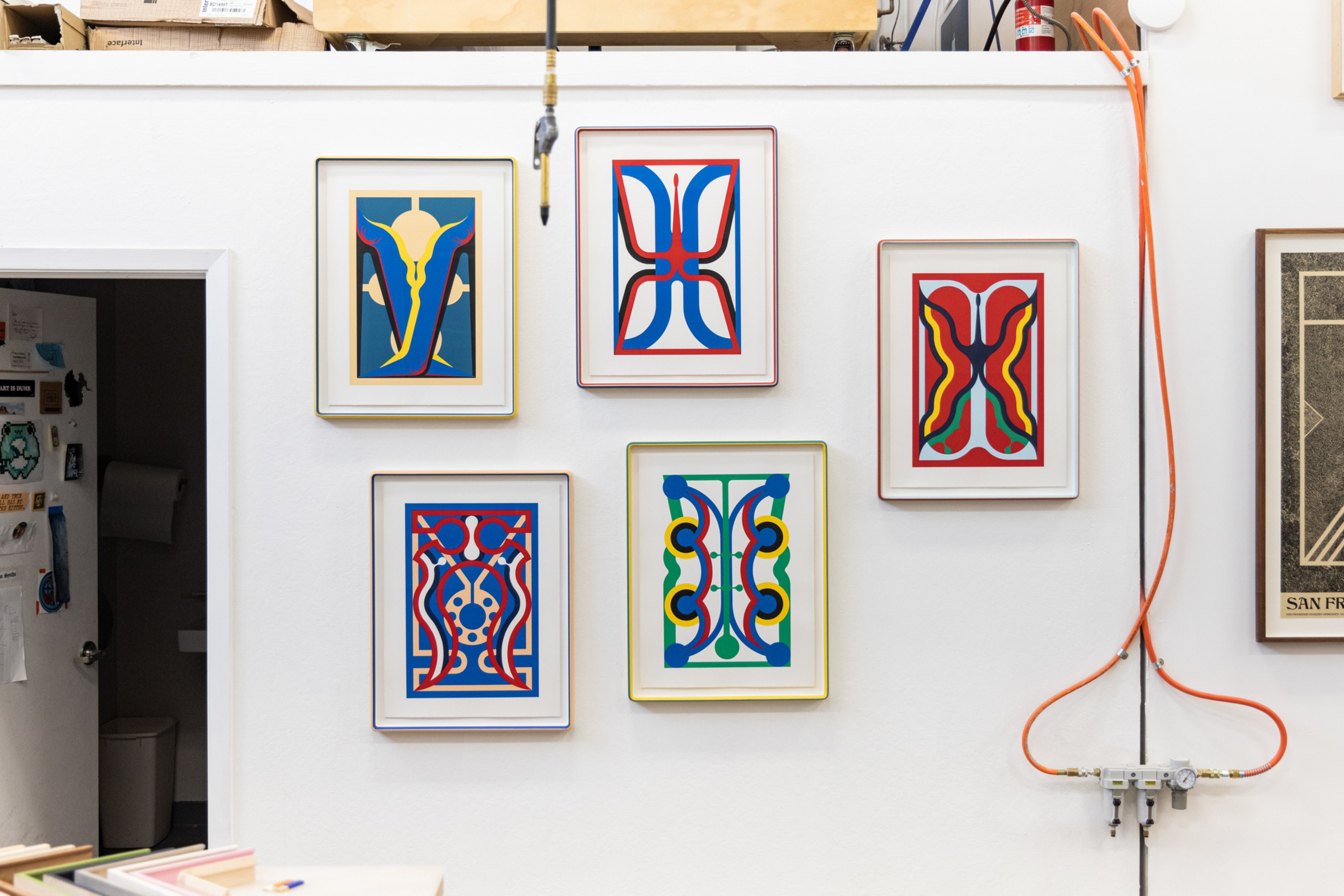 Five colorful abstract geometric artworks with symmetrical designs hang on a white wall, with a partially open door and visible orange cables nearby.
