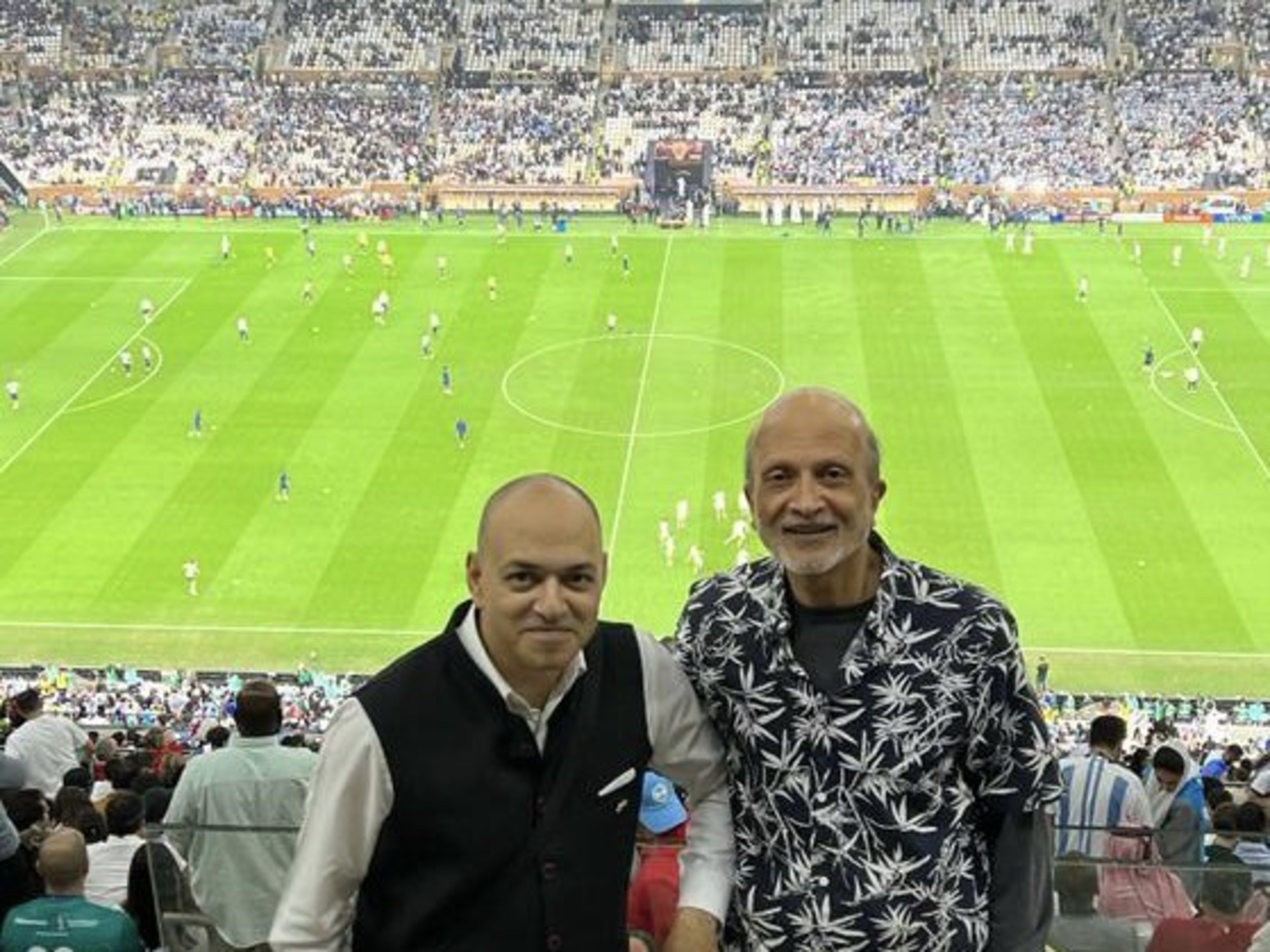 Two men pose in front of a soccer field during a match, with players on the field and thousands of spectators filling the stadium seats behind them.