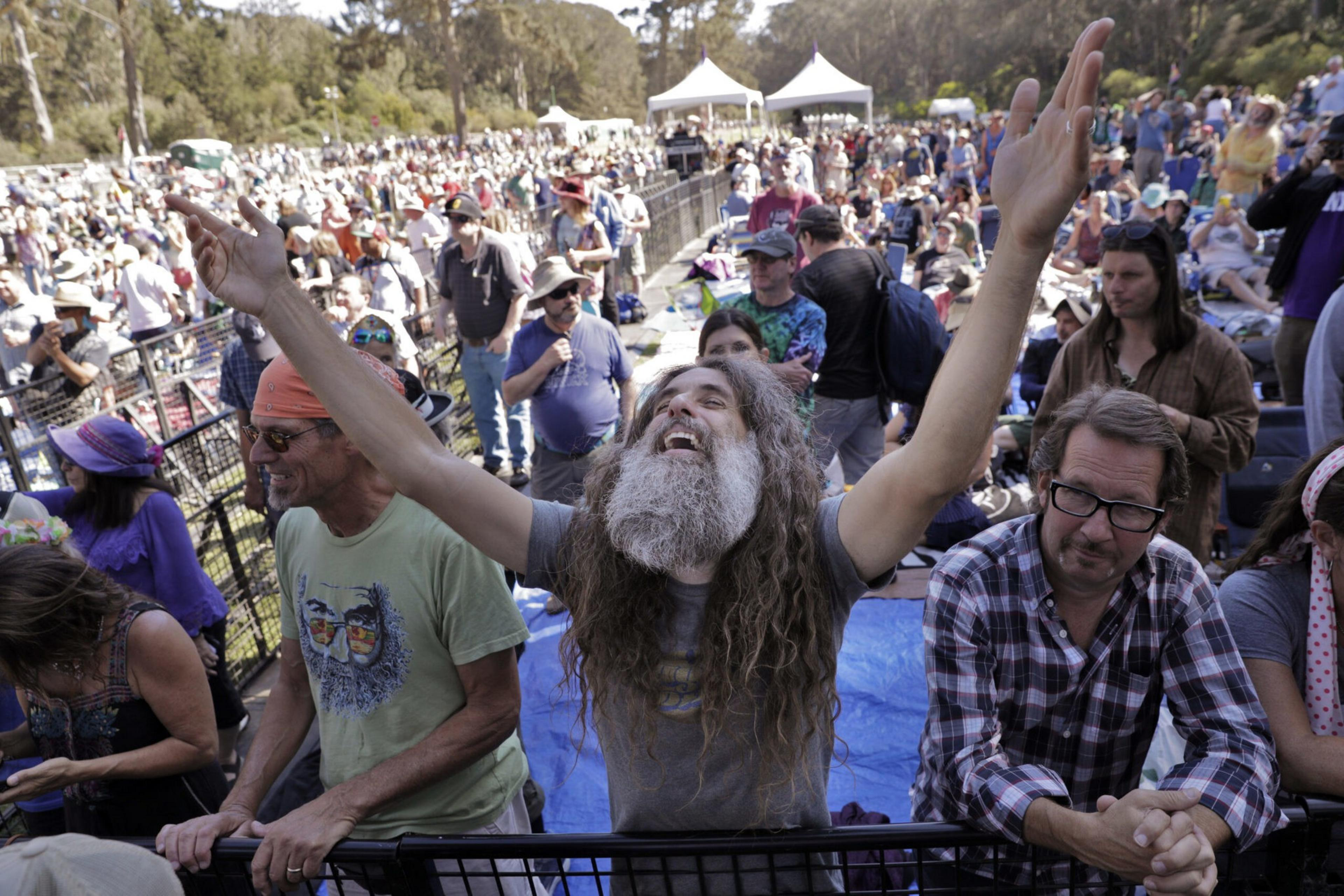 A crowd of people at an outdoor event, with a bearded man in the center raising his arms joyfully while others around him watch or relax.