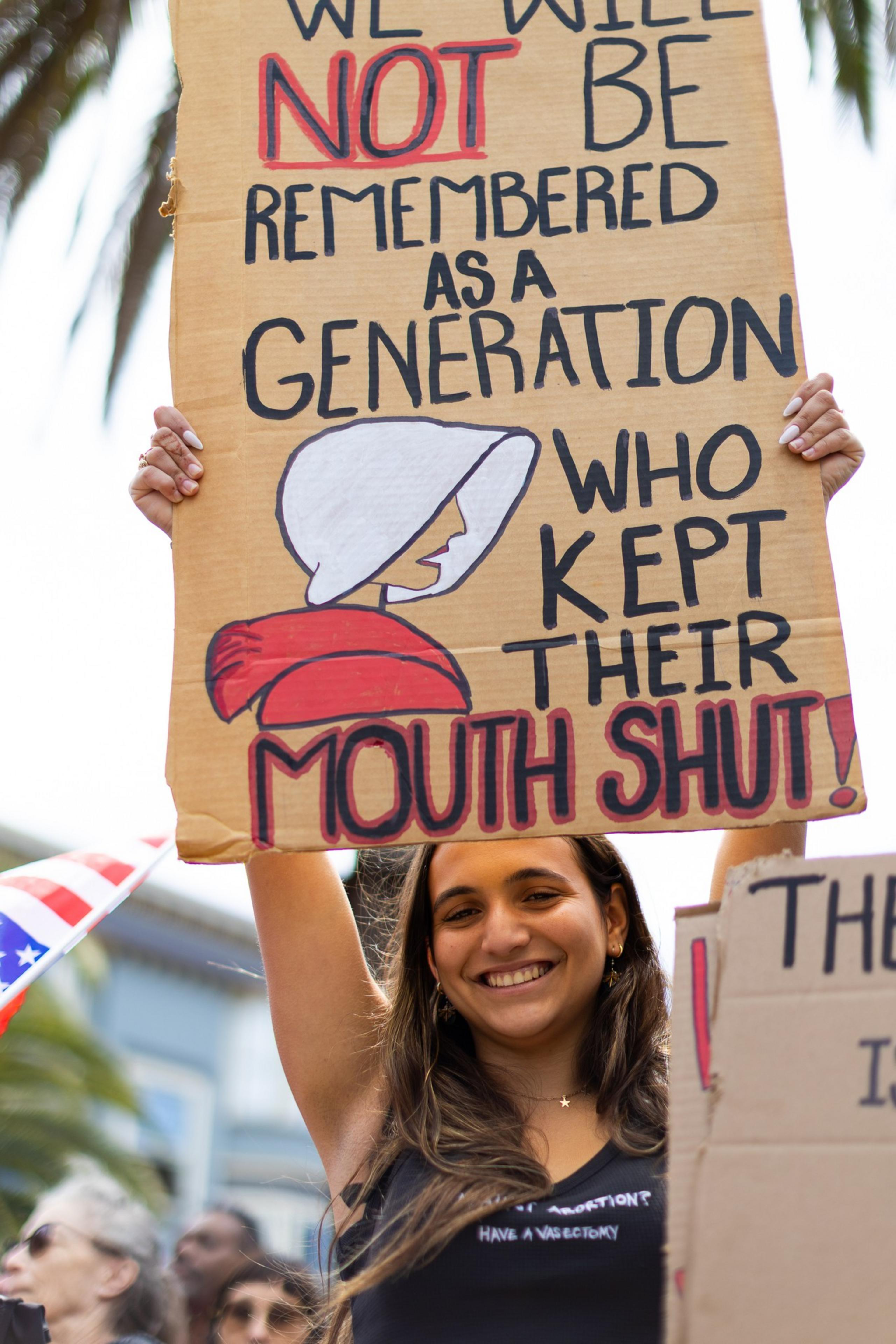 A smiling woman holds up a sign with bold text challenging silence, featuring an illustration of a woman in a white bonnet. People and a flag are in the background.