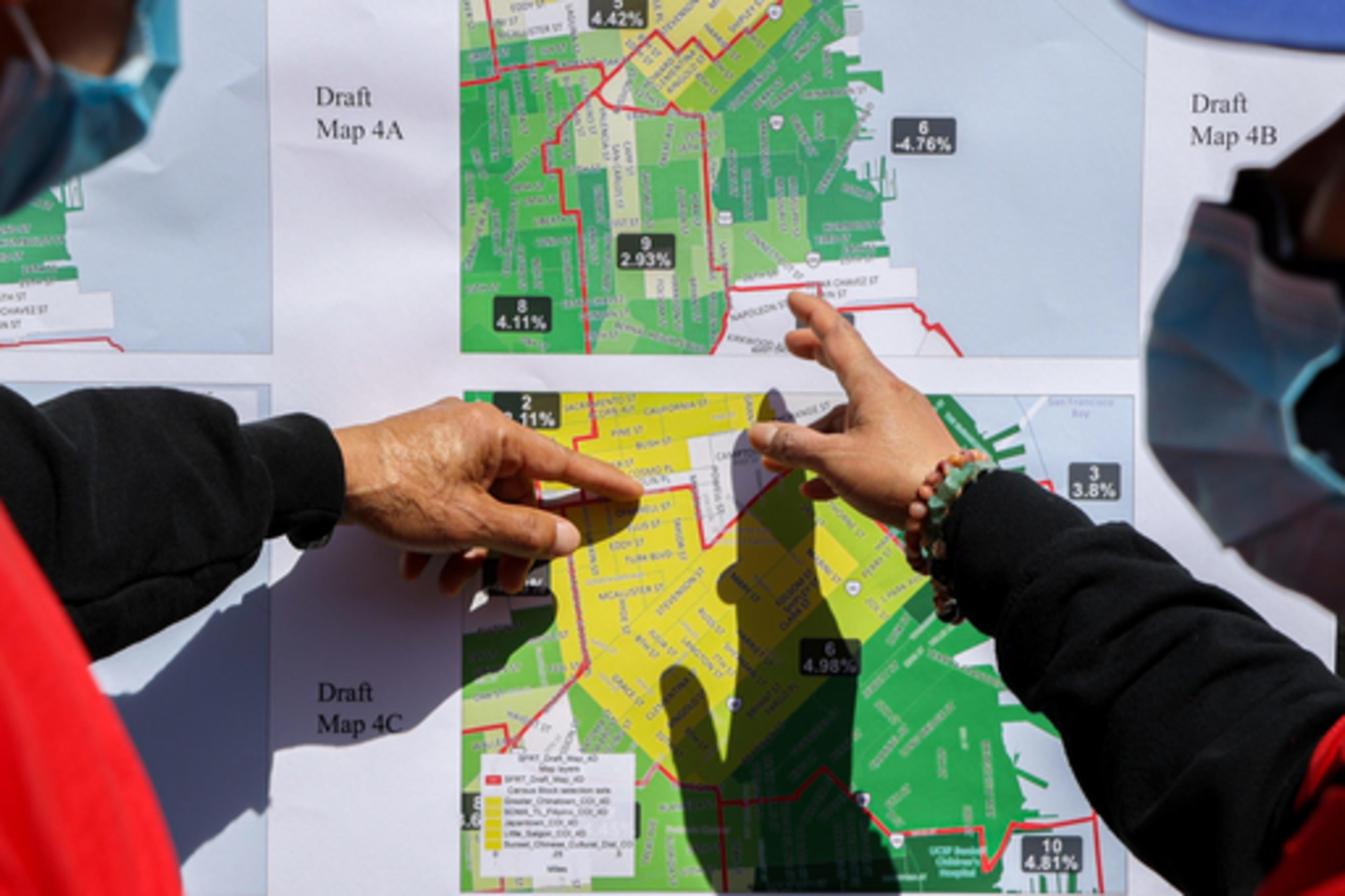 Two people wearing masks point at colored draft maps labeled 4A, 4B, and 4C, showing different zones and percentages within city boundaries.