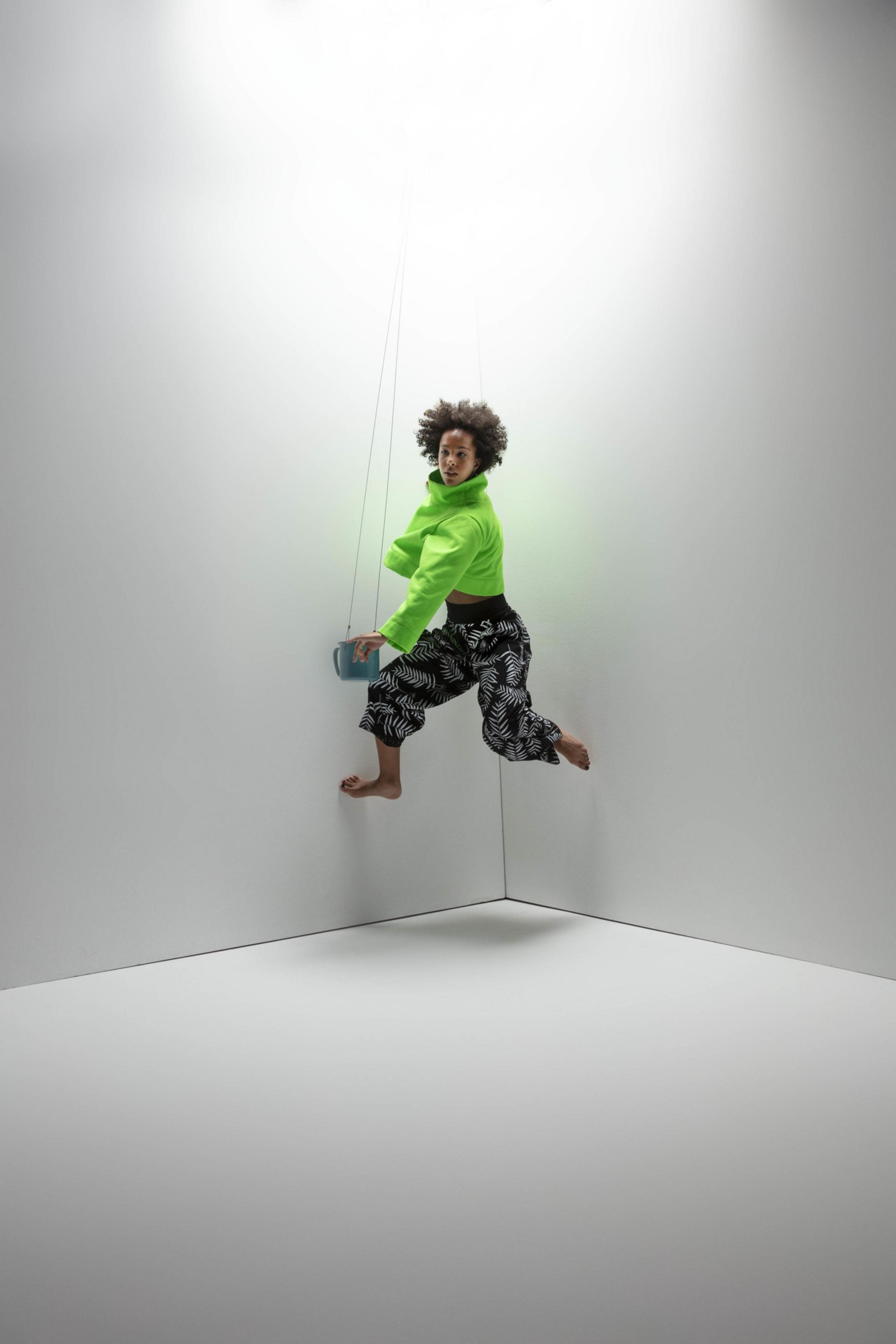 A person in a bright green jacket and patterned black-and-white pants appears to float mid-air in a white corner, holding a small blue radio.