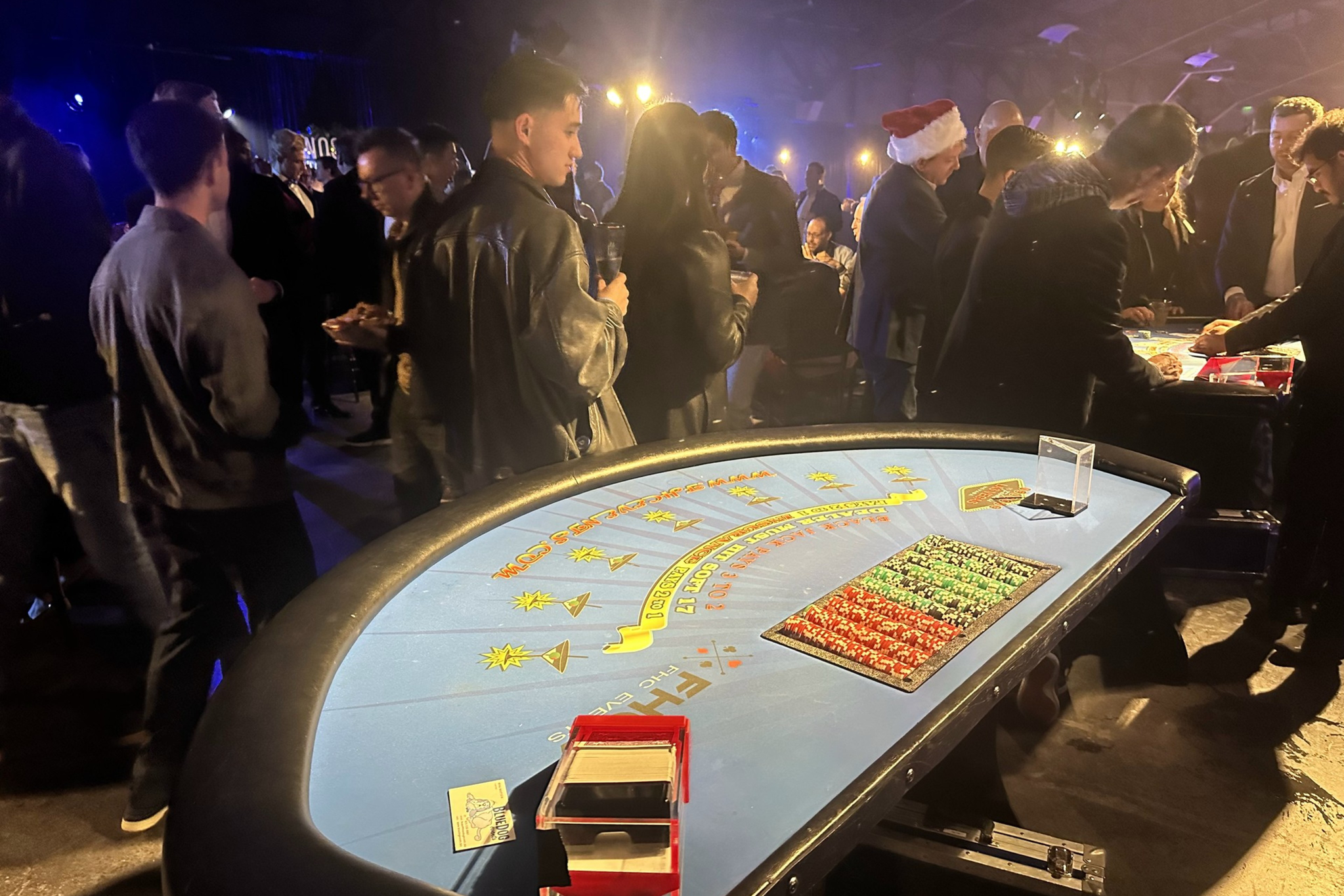 People are gathered around casino tables in a dimly lit room. Some are wearing festive attire, and there are chips and cards on the tables.