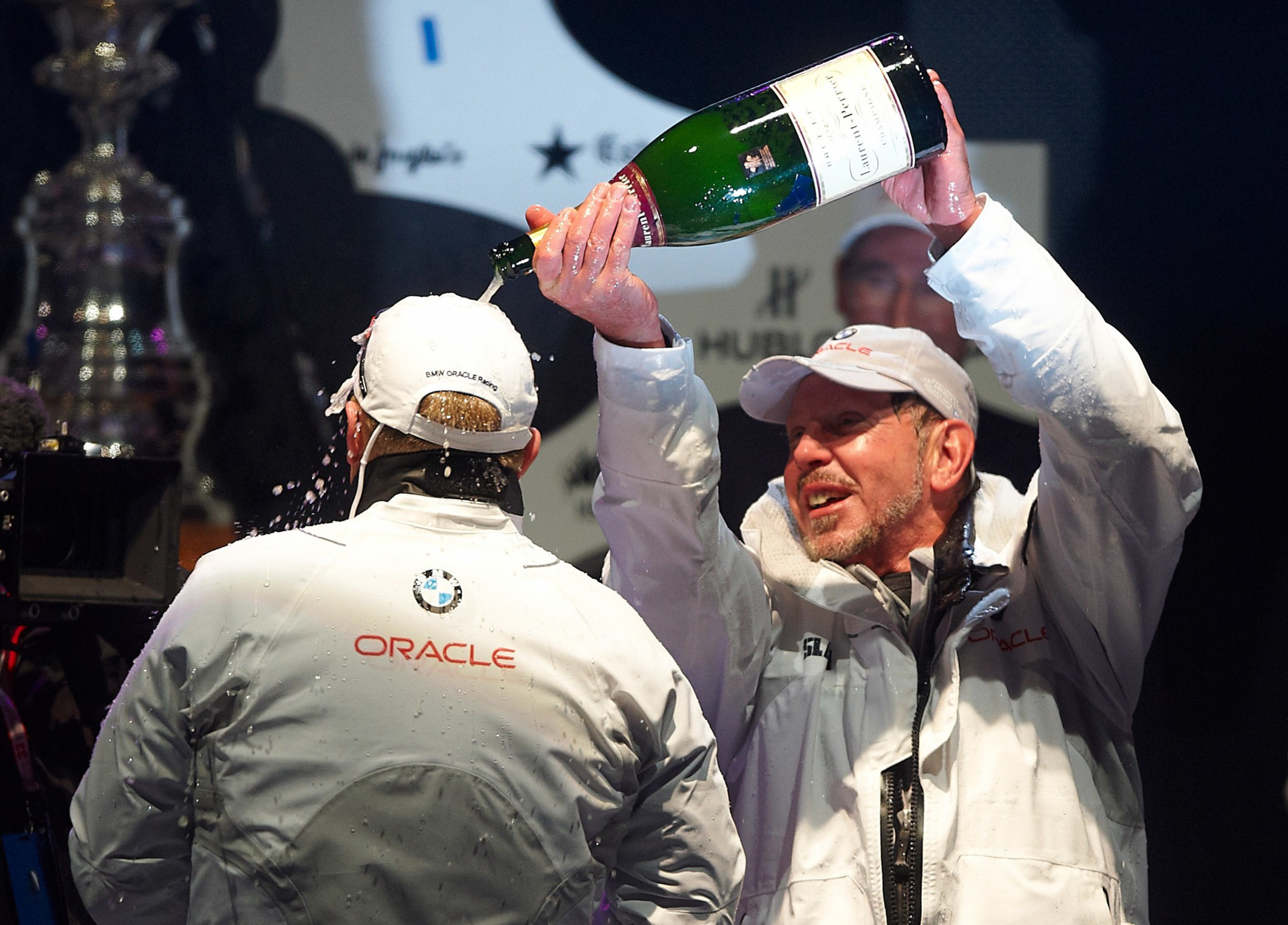 A man in white Oracle gear pours champagne over the head of another man in similar clothing during a celebration.