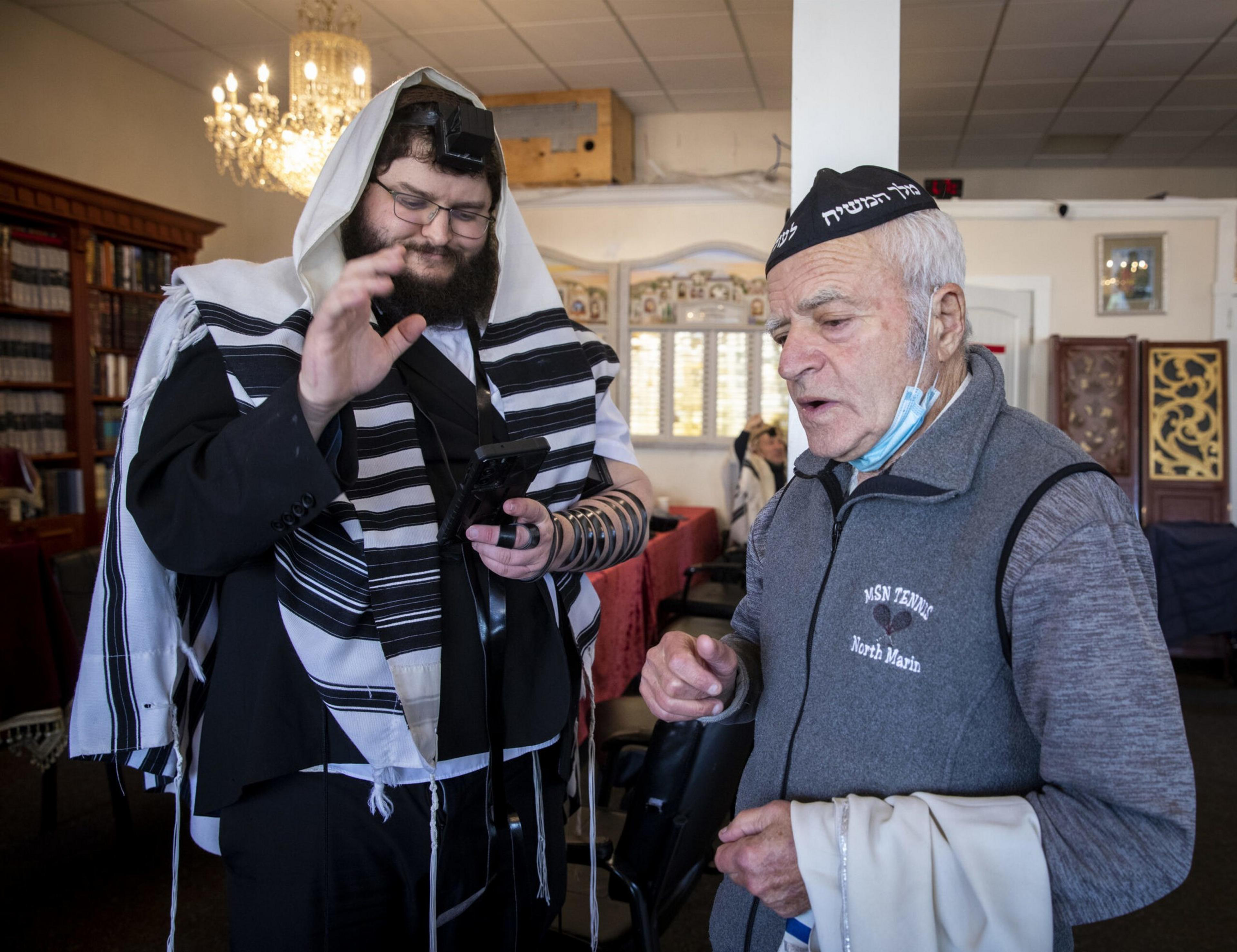 A man in traditional Jewish prayer attire and tefillin uses a smartphone while talking to an older man wearing a kippah and face mask under his chin.