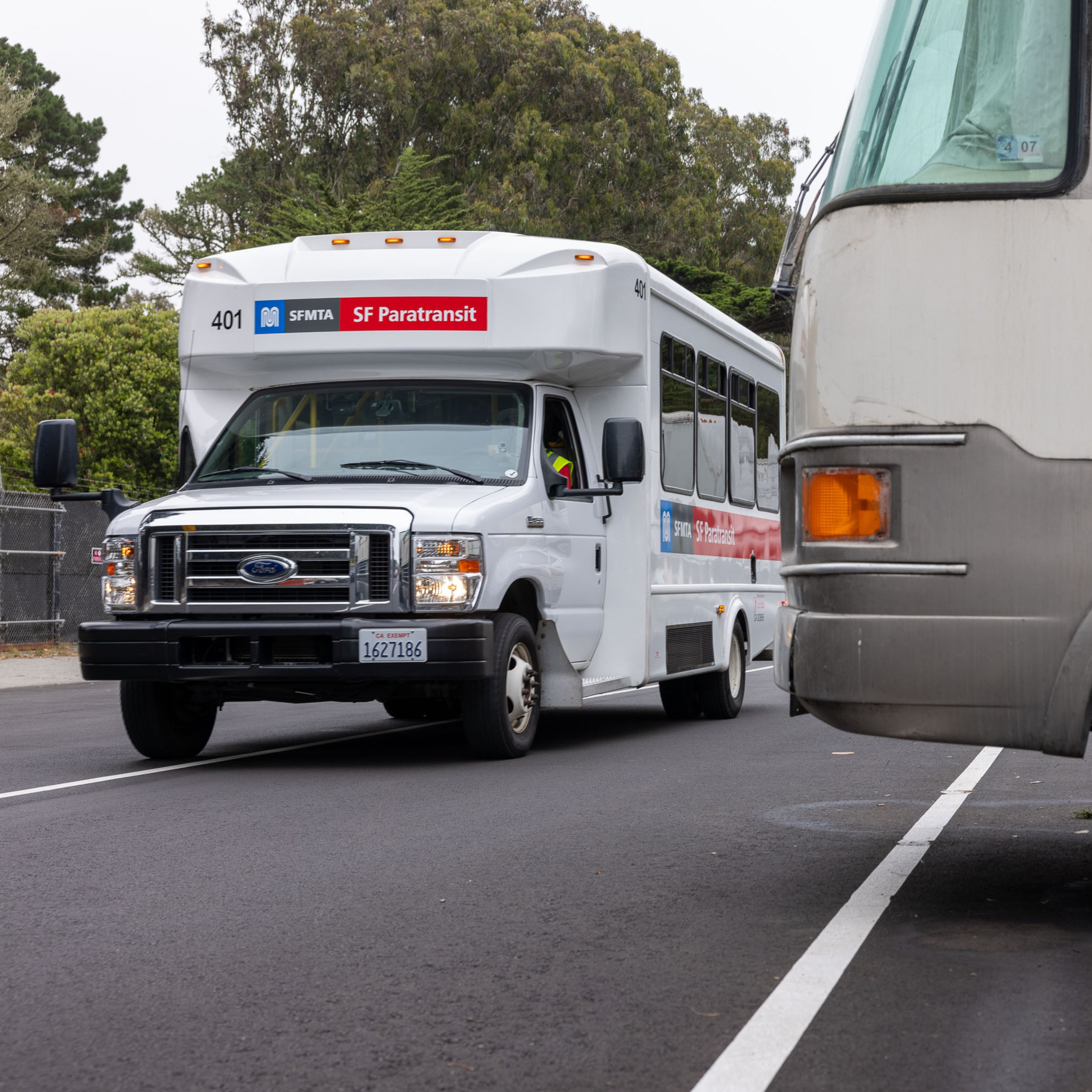 A white SF Paratransit bus numbered 401 is traveling on a paved road, passing by a parked vehicle on the right side, with trees in the background.