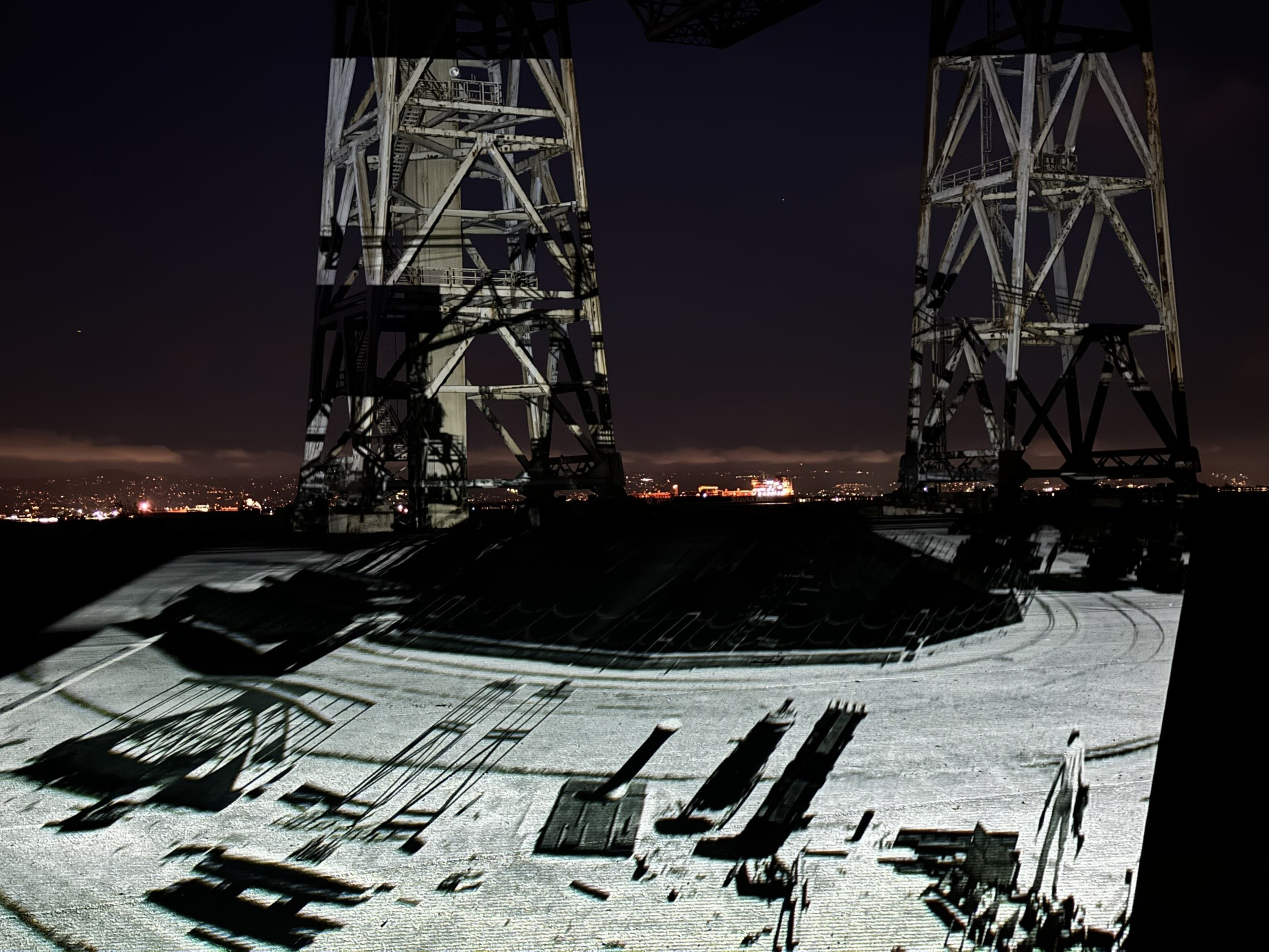 A night scene with a large metal structure and projected images on the ground. In the distance, city lights and a ship are visible against a dark sky.